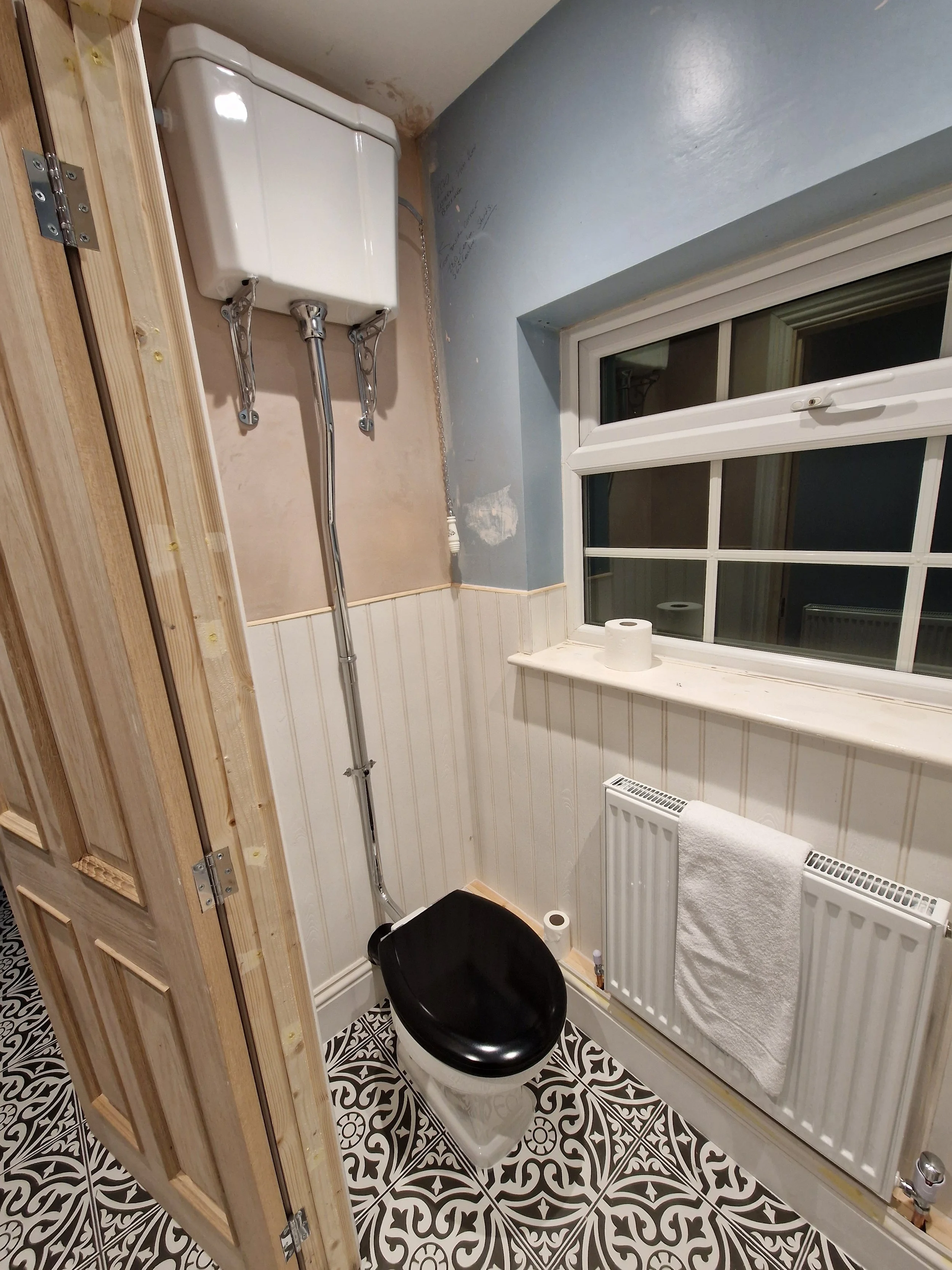 A small bathroom under renovation with a black toilet seat, a window, a radiator with a towel, an open wall with exposed wooden framing, and a white tank mounted high on the wall near the ceiling.
