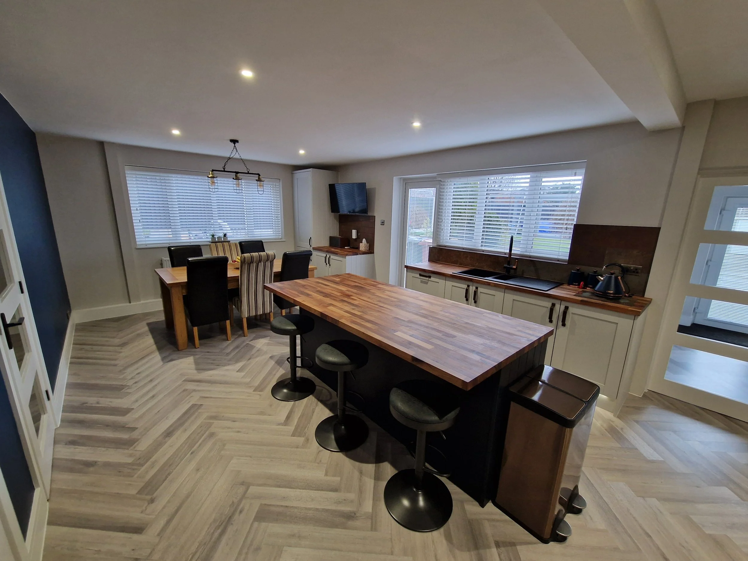 Modern kitchen and dining area with wooden countertops, black bar stools, dining table, and large windows with blinds.