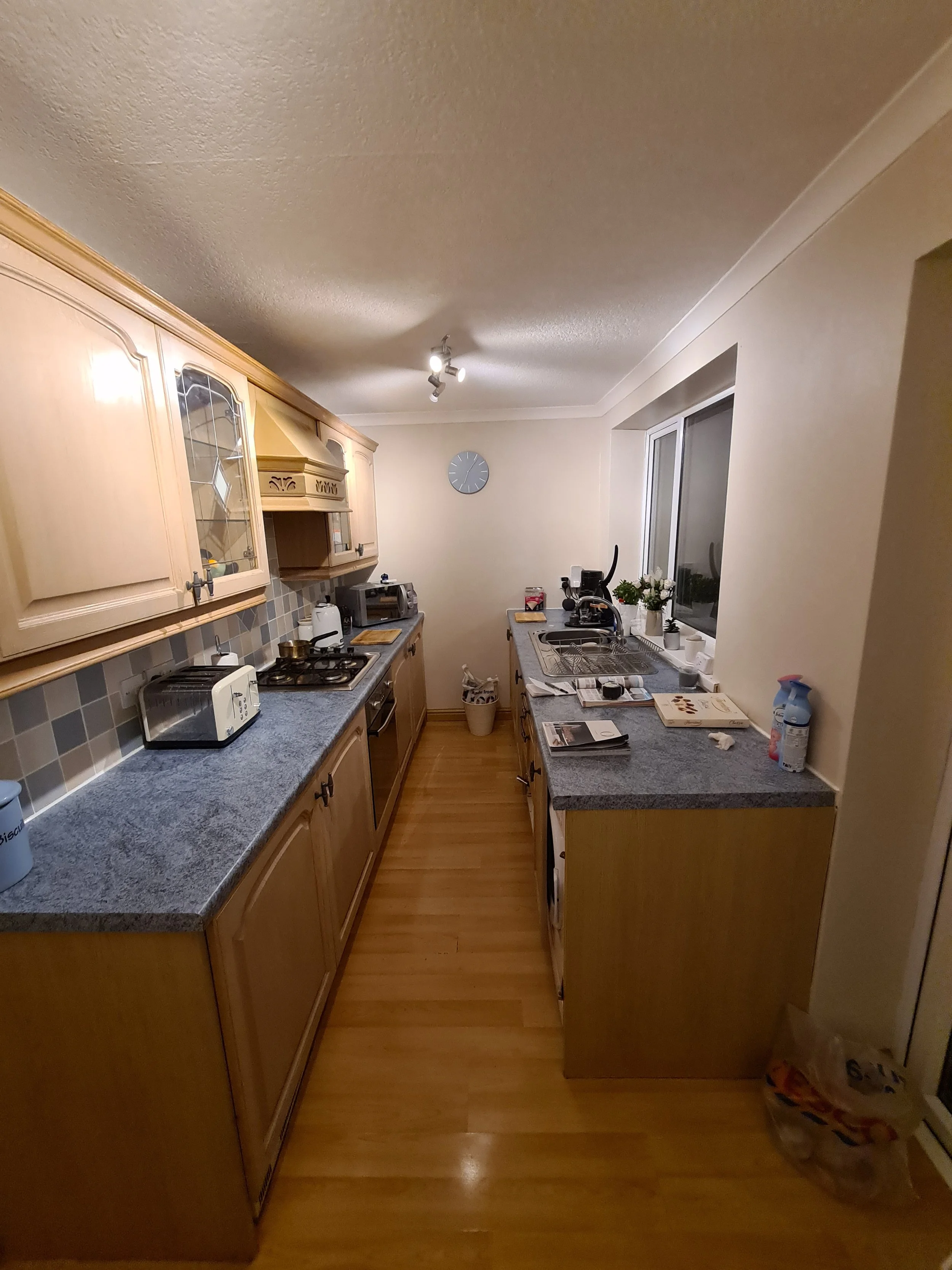 A narrow kitchen with wooden cabinets, a blue granite countertop, and a window above the sink. Items on the countertops include a toaster, microwave, plant, and cleaning spray. A clock on the wall and wooden flooring are visible.