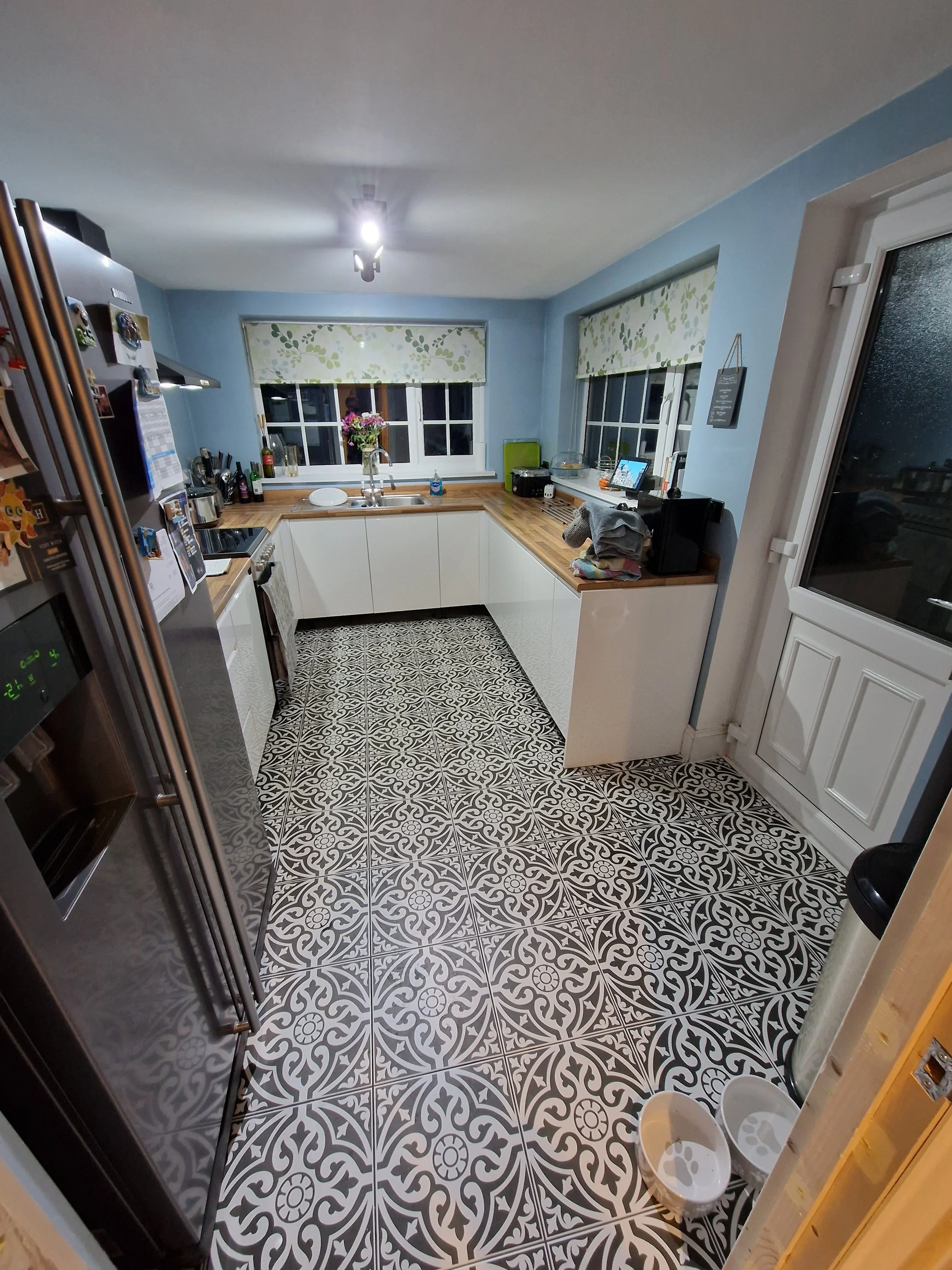 Kitchen with patterned black and white floor, blue walls, white cabinets, and windows with floral blinds. Items on the counter include a vase with flowers, bottles, and small appliances.