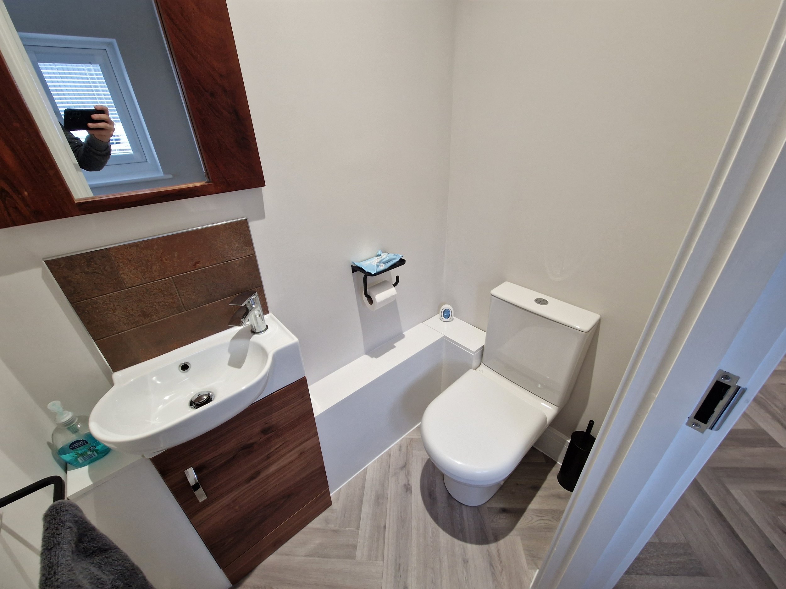 Small bathroom with a white toilet, a small sink with a silver faucet, a wooden cabinet beneath the sink, a mirror above the sink, a window reflected in the mirror, a black toilet brush holder, a container of hand soap, a small shelf with tissue and a clock, white walls, and wood-patterned floor.