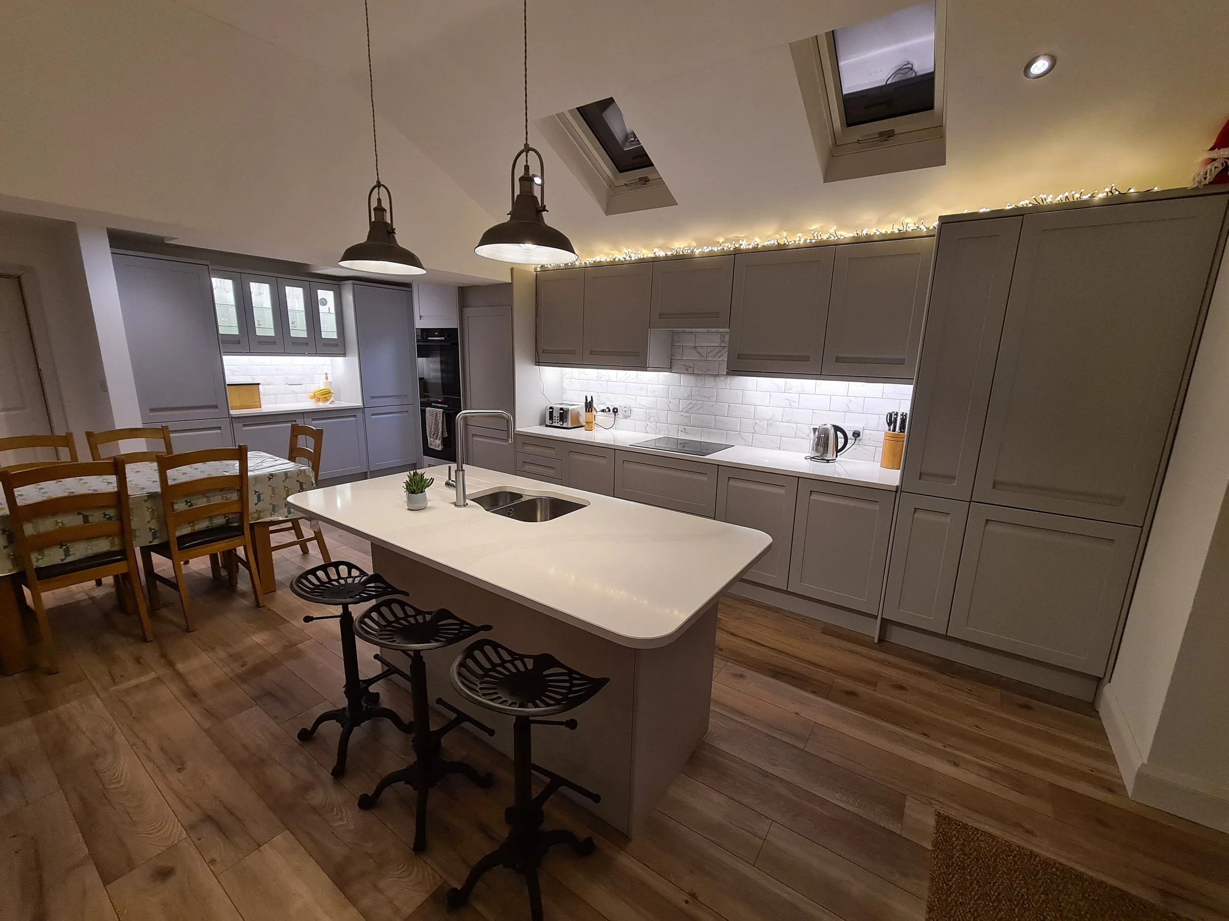 Modern kitchen with gray cabinets, white marble backsplash, and an island with a white countertop. There are three black bar stools at the island, a dining table with wooden chairs, and pendant lights hanging from the ceiling. The kitchen has skylights and warm lighting.