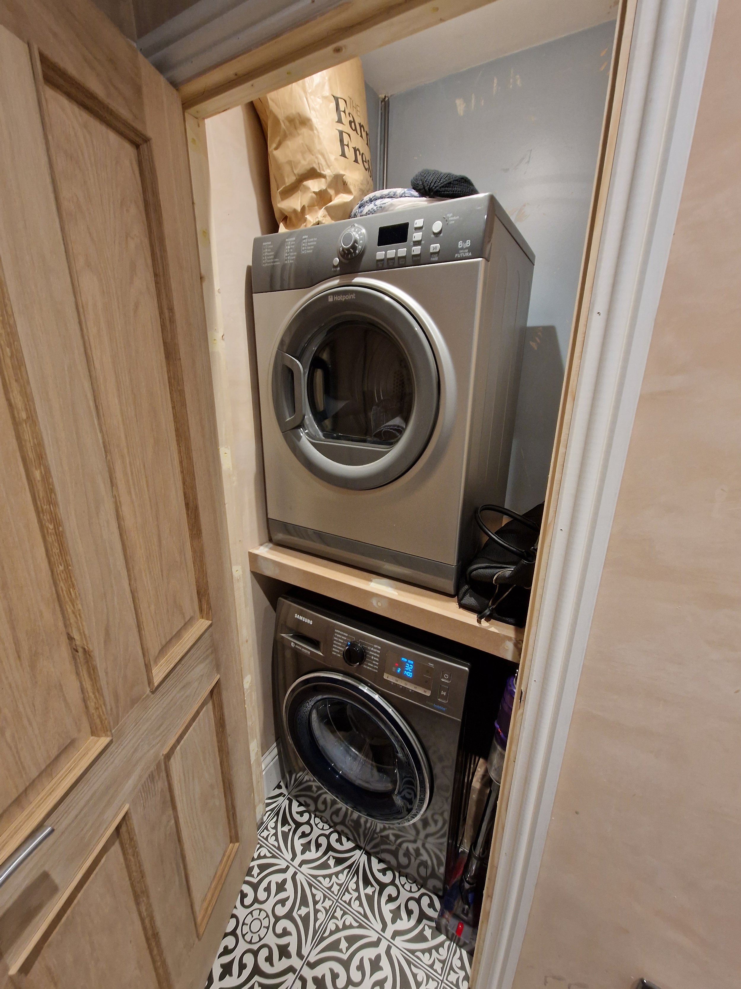A small laundry room with a stacked washer and dryer, wooden shelving, and patterned tile floor.