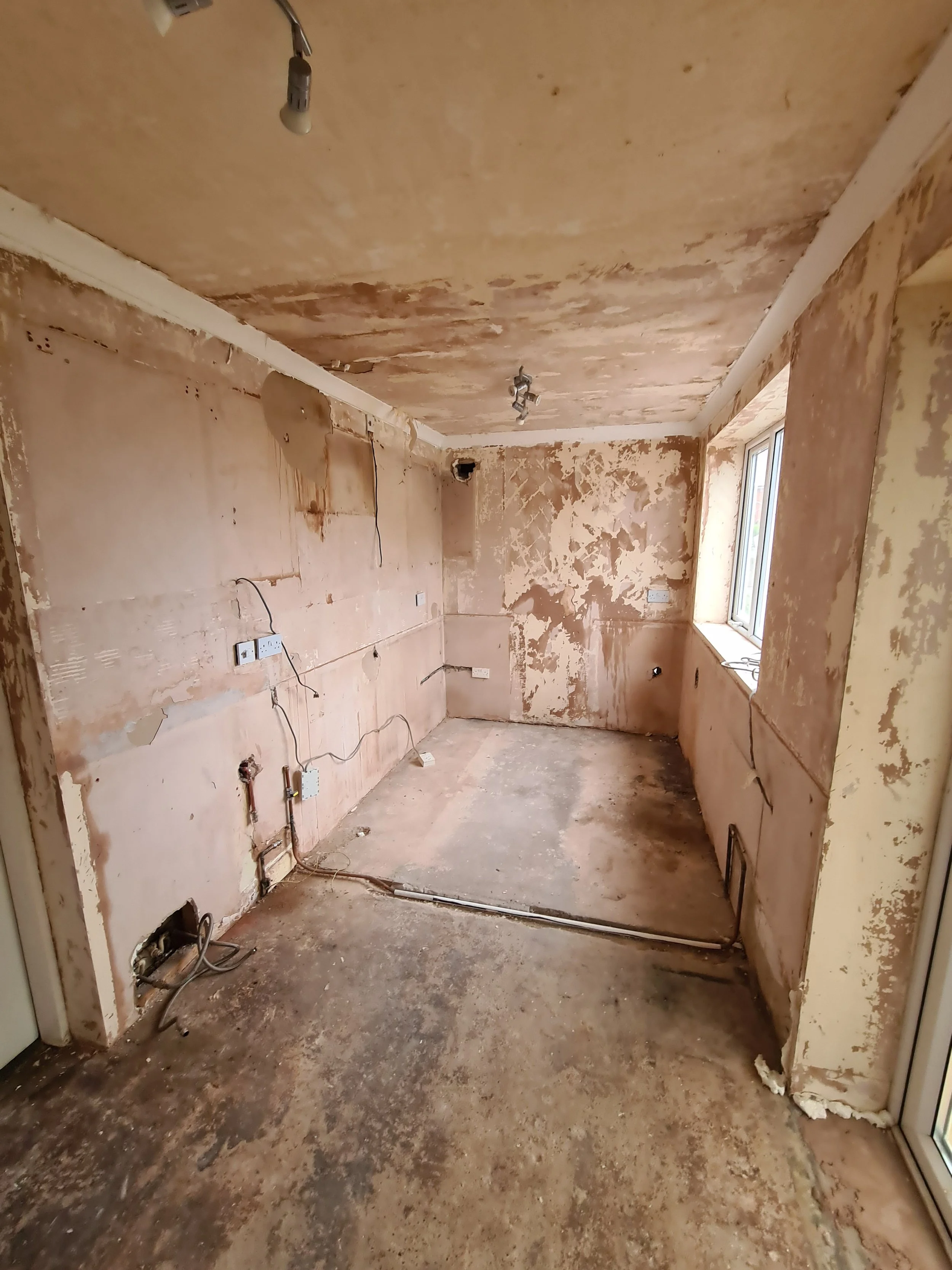 Empty kitchen with peeling paint, exposed wiring, and unfinished walls and floor under renovation.