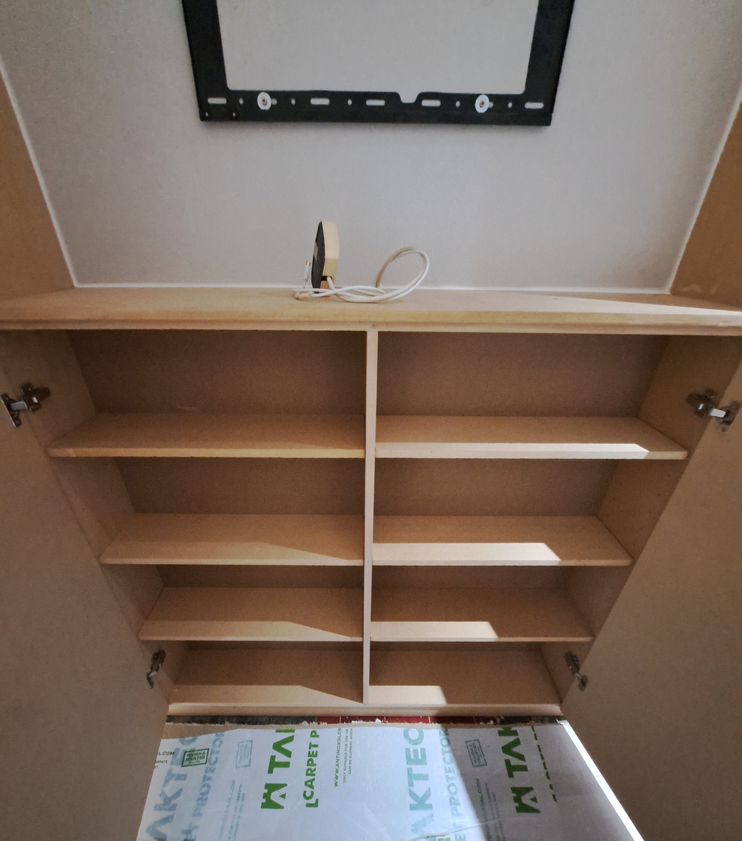 Empty wooden cabinet with four shelves and two open doors, mounting bracket on top shelf, electrical cord and device above cabinet, floor covered with protective sheet.