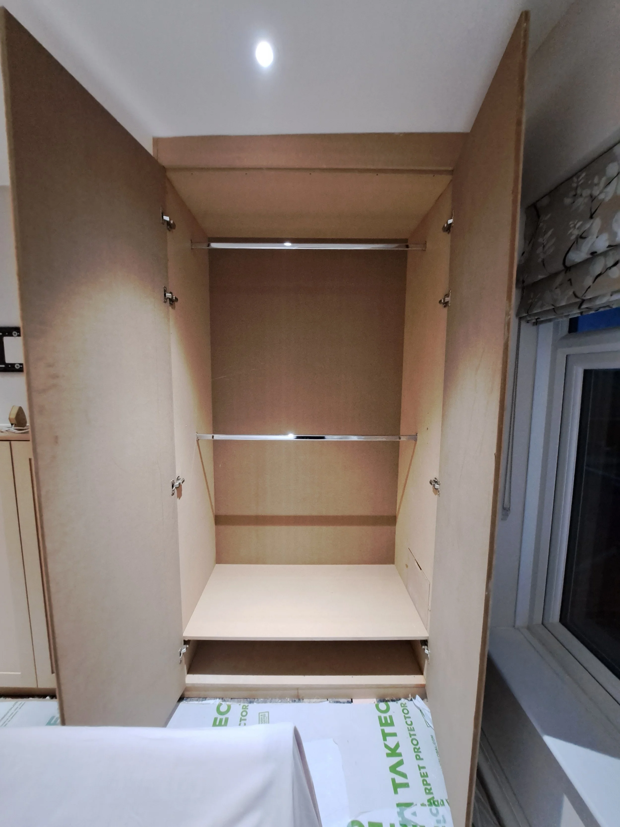 Empty wooden wardrobe with open doors and metal rods for hanging clothes, situated near a window with floral curtains.