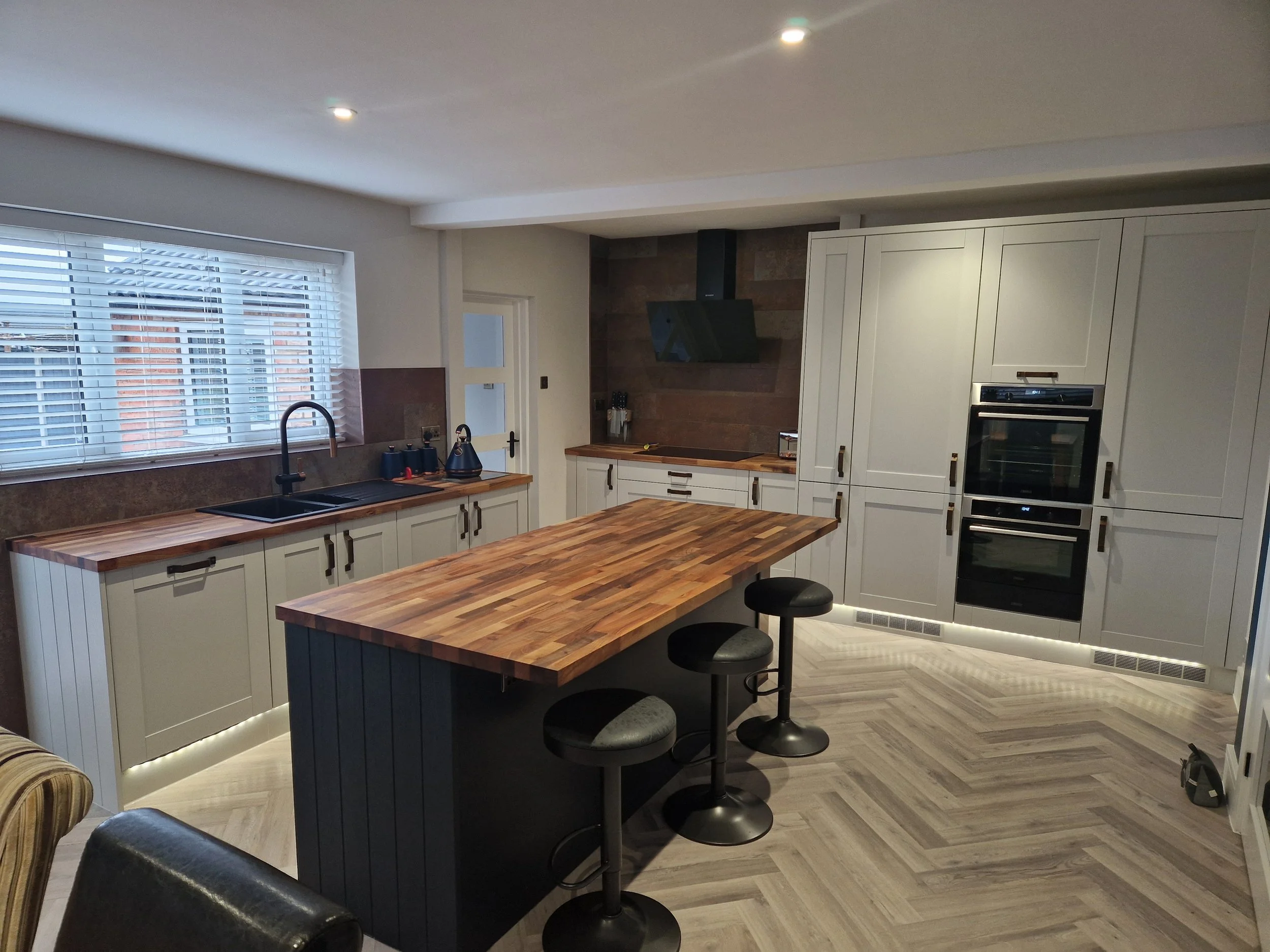 Modern kitchen with white cabinets, wooden countertops, a central island with a wooden top and black stools, a black oven, and a window with blinds.