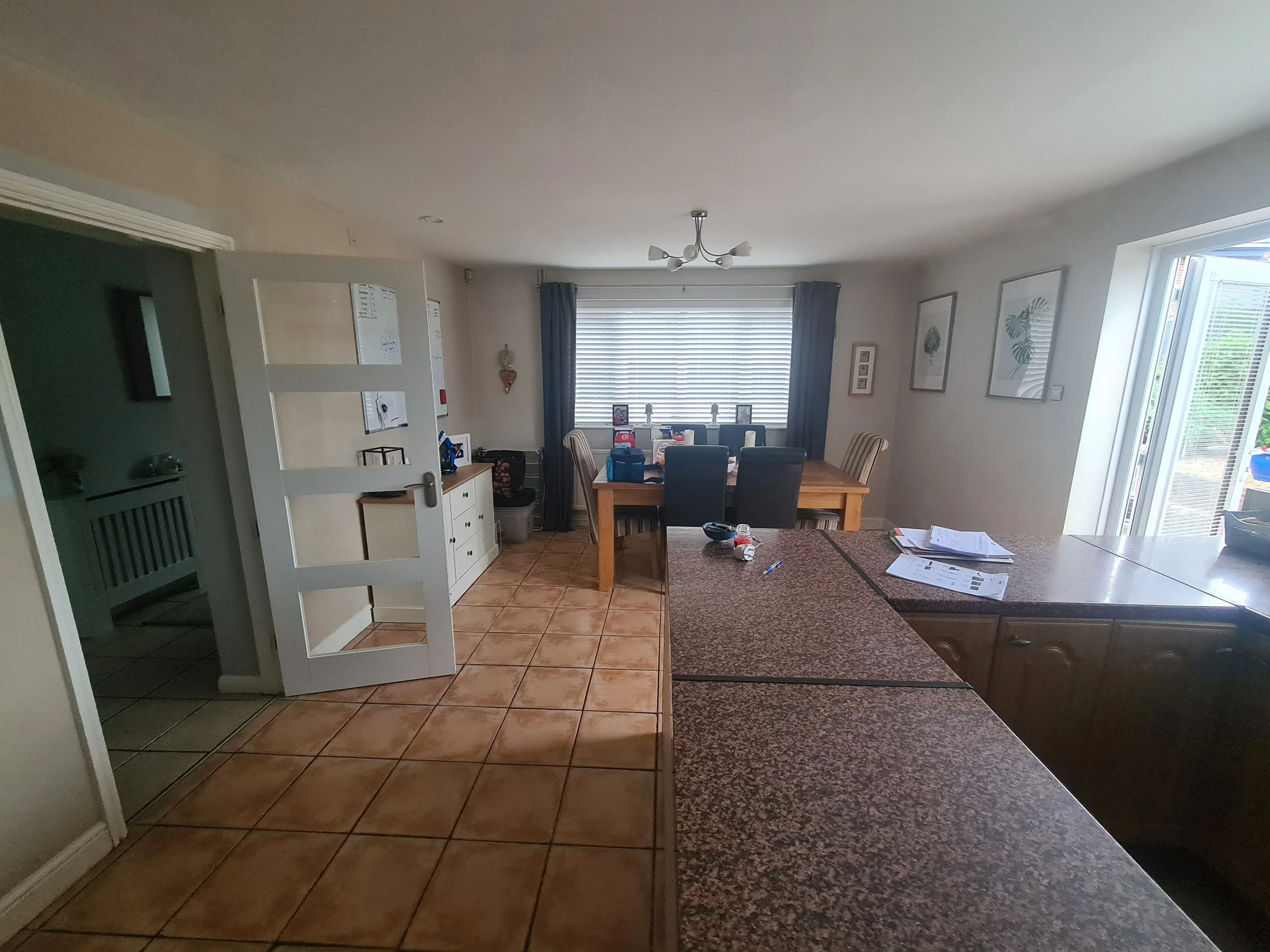 Dining area with a wooden table and six chairs, window with blinds and curtains, white walls with framed artwork, adjacent kitchen with granite countertops, and a tiled floor.