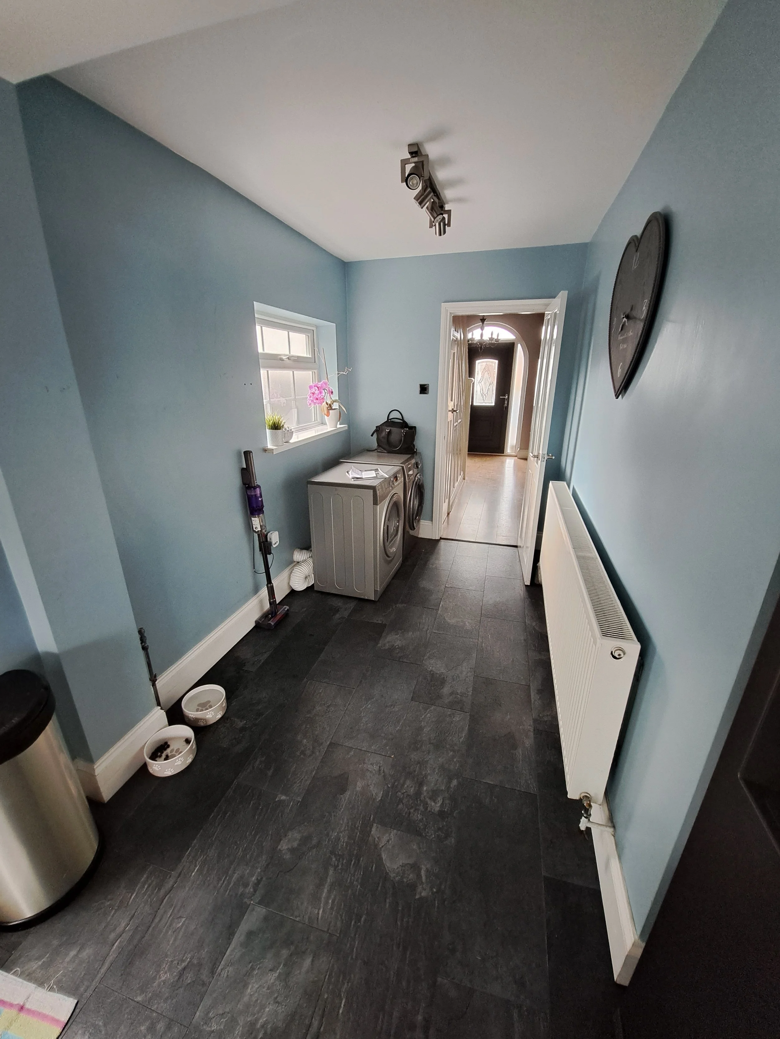 Laundry room with light blue walls, black tile flooring, a window with potted plants, a washing machine and dryer, a black handbag on top, a vacuum cleaner, a wall clock, a radiator, and an open door leading to a hallway with a front door visible in the background.