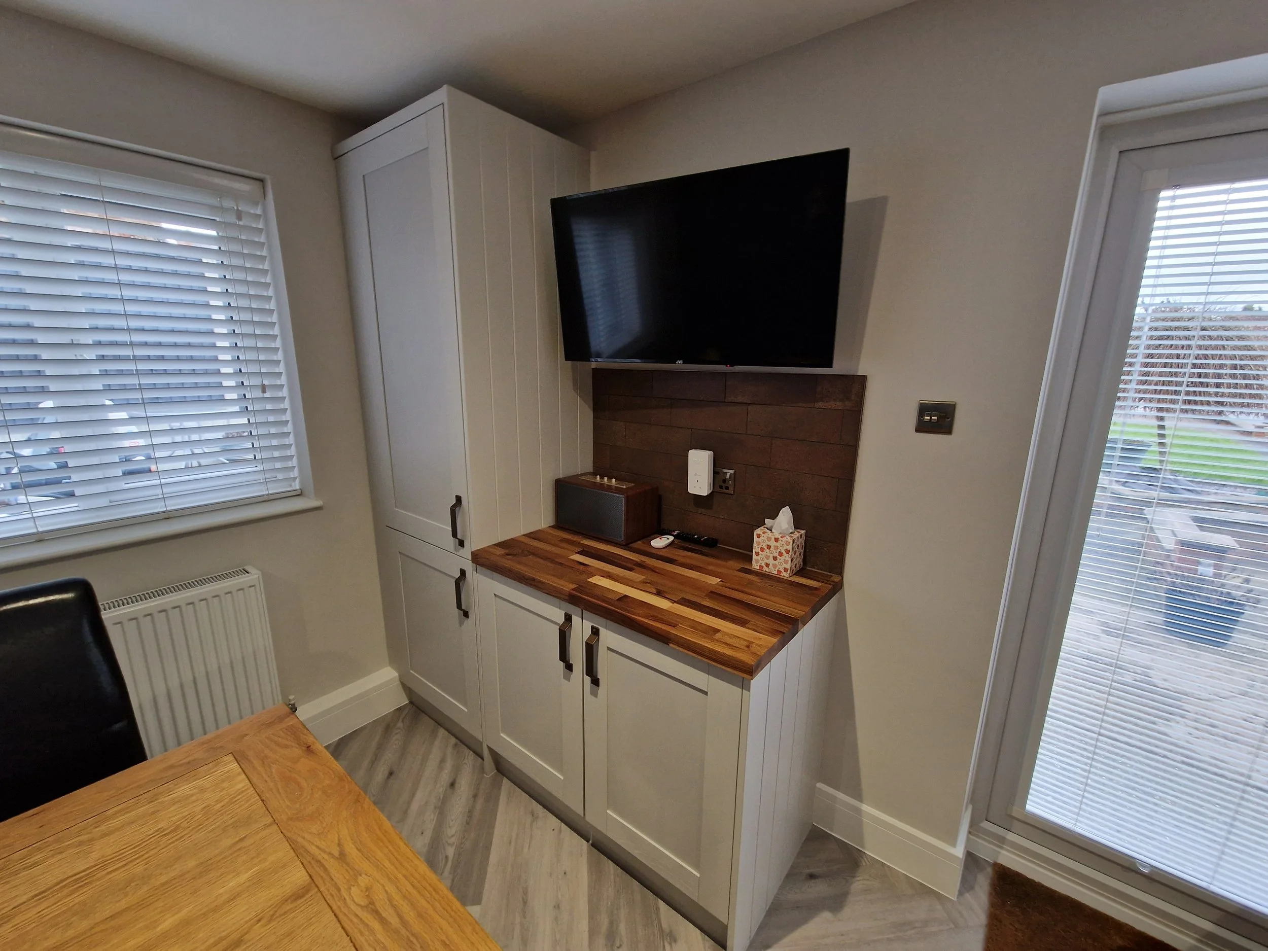 Interior of a room showing a white cabinet with a dark wood countertop, a flat-screen TV mounted on the wall, two windows with blinds, and part of a wooden table.