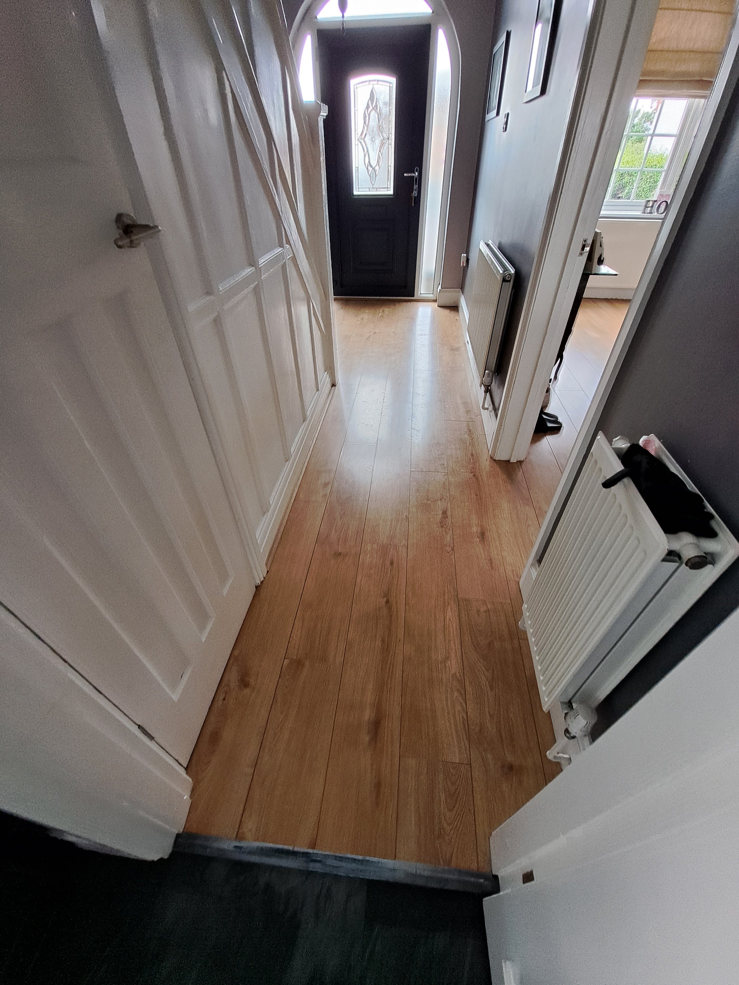 Interior view of a hallway with wooden flooring and a black front door with a decorative glass panel, with part of a room visible on the right side and a white radiator on the right wall.