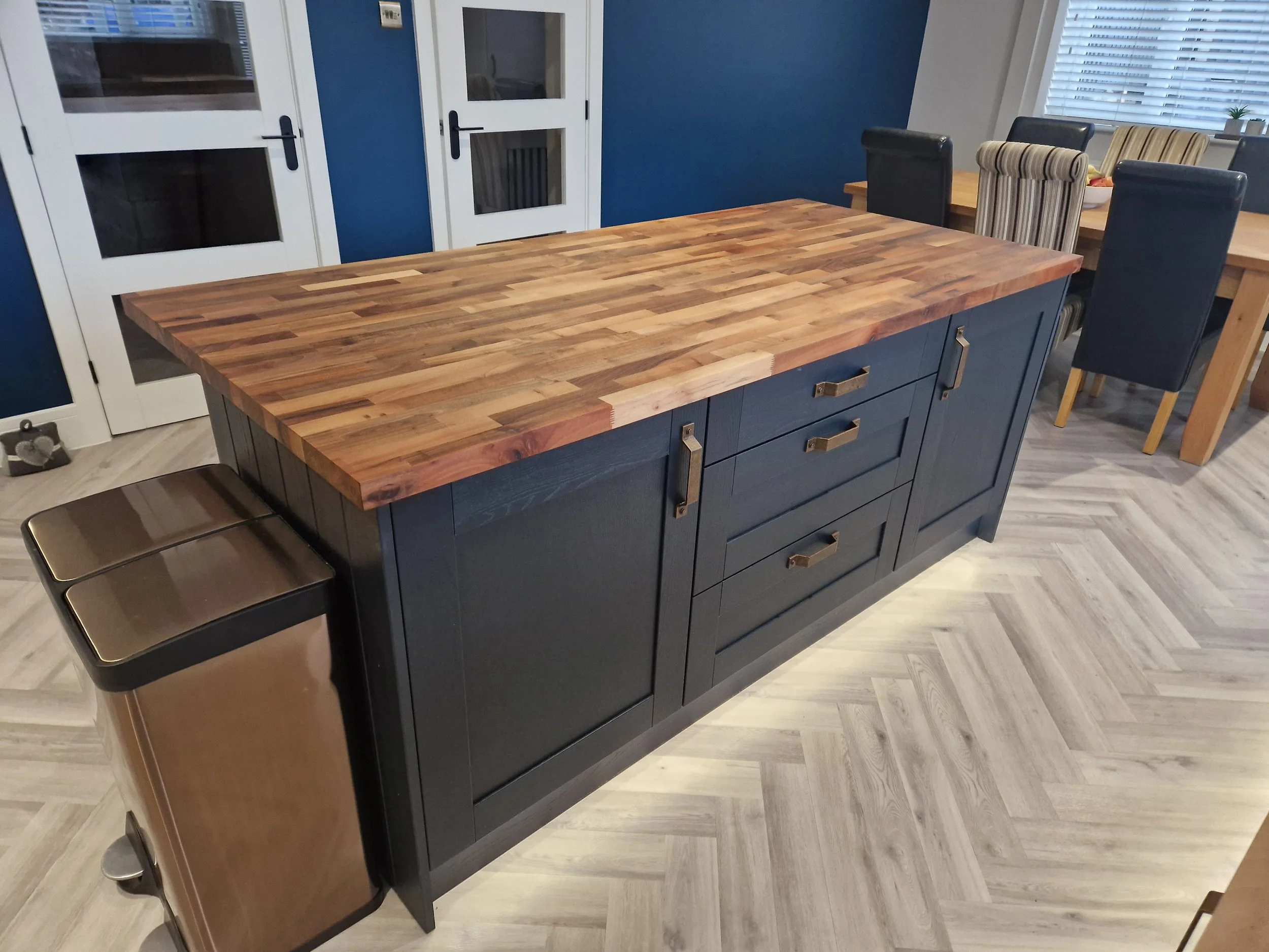 A kitchen island with a wooden butcher block countertop, navy blue base, and metal handles, next to a brown trash bin. In the background, there is a dining table with chairs and white doors with glass panels.
