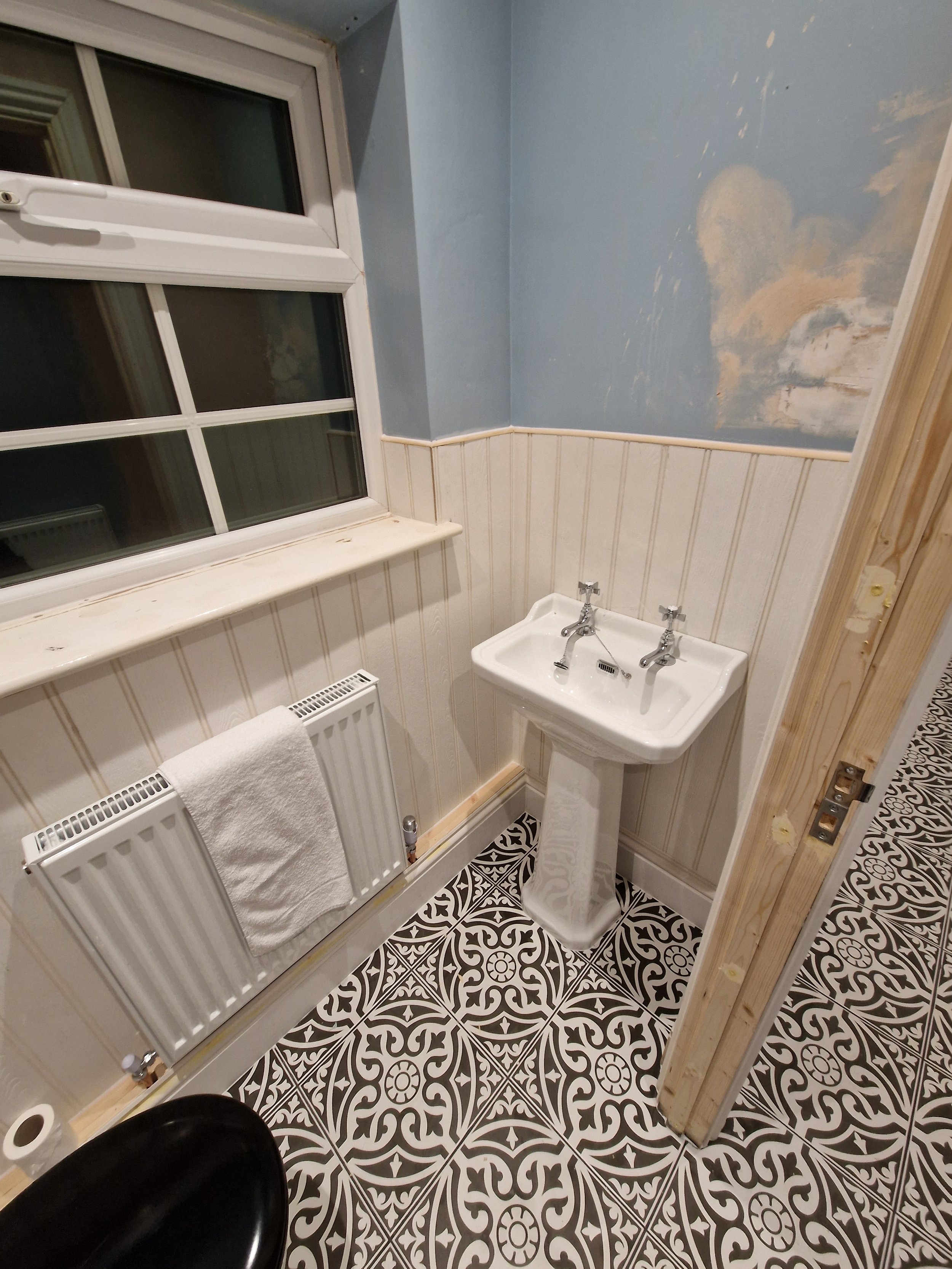 Small bathroom with patterned black and white floor tiles, white sink, radiator with towel, window, and partially painted wall.