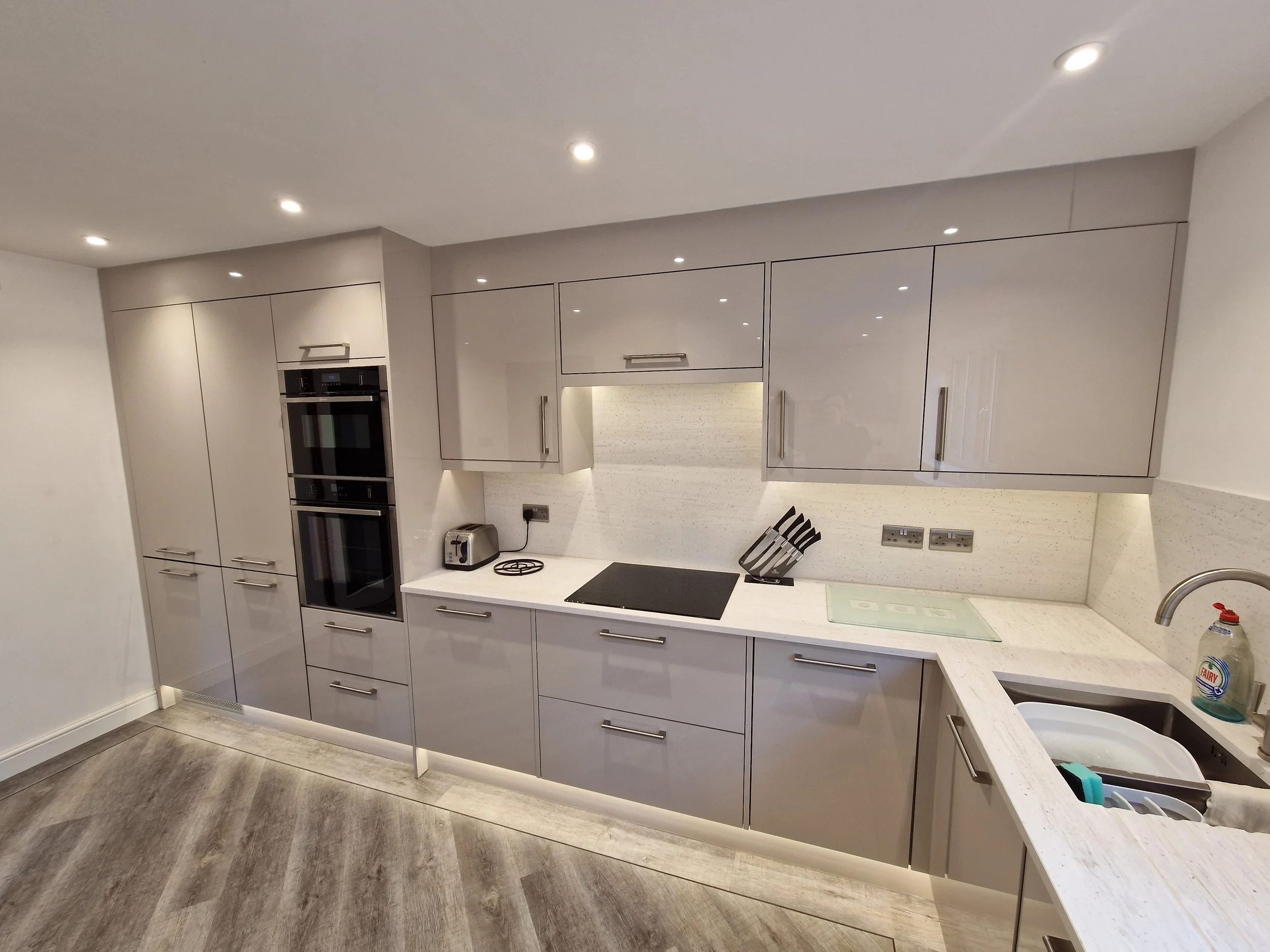 Modern kitchen with grey cabinets, built-in oven, toaster, induction cooktop, knife block, and sink with dish soap, illuminated by ceiling lights.
