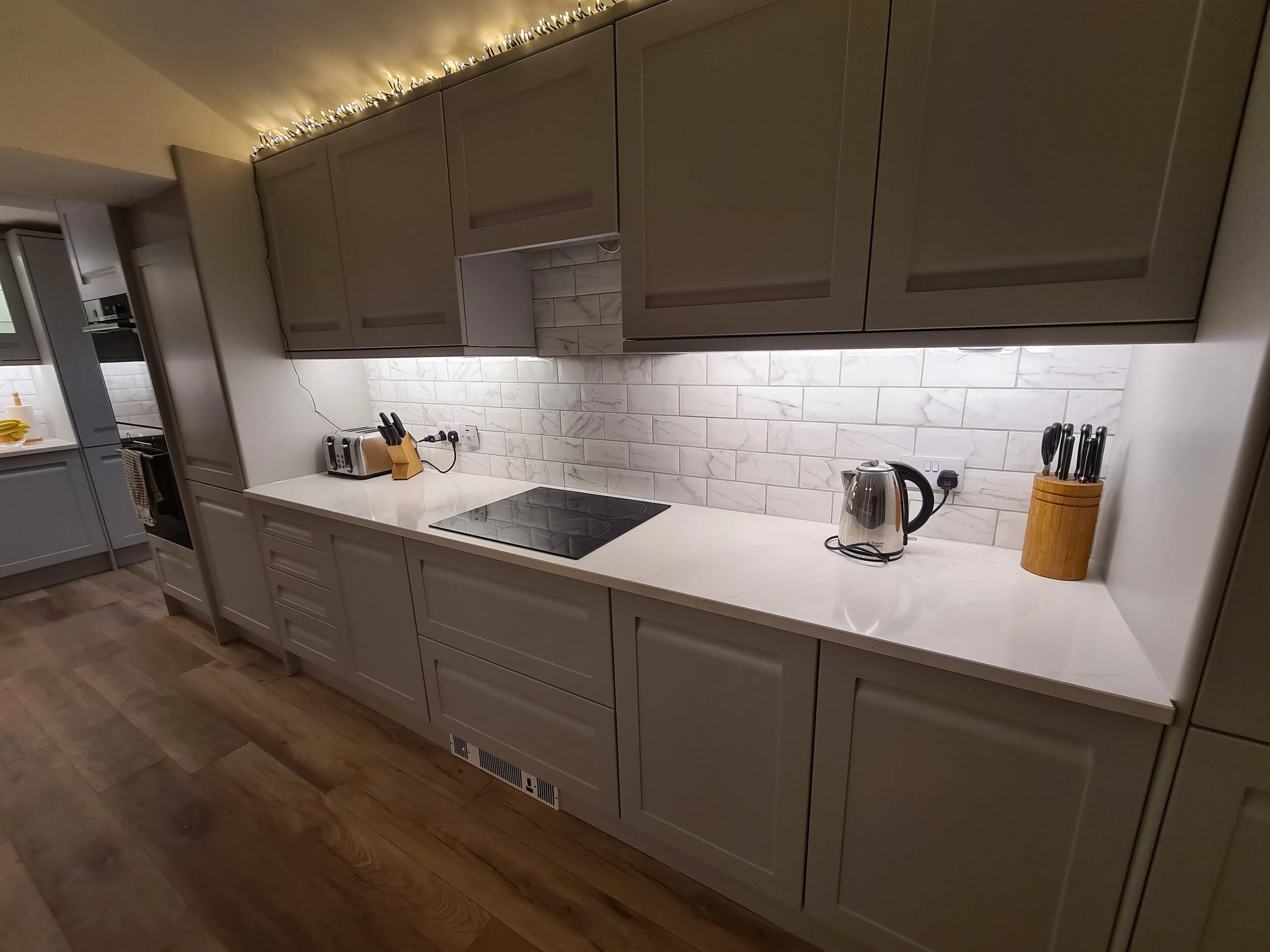 Modern kitchen with white cabinetry, marble backsplash, and wooden flooring. Countertop has a toaster, a knife block, an electric kettle, and electrical outlets. Overhead lighting and decorative string lights above cabinets.