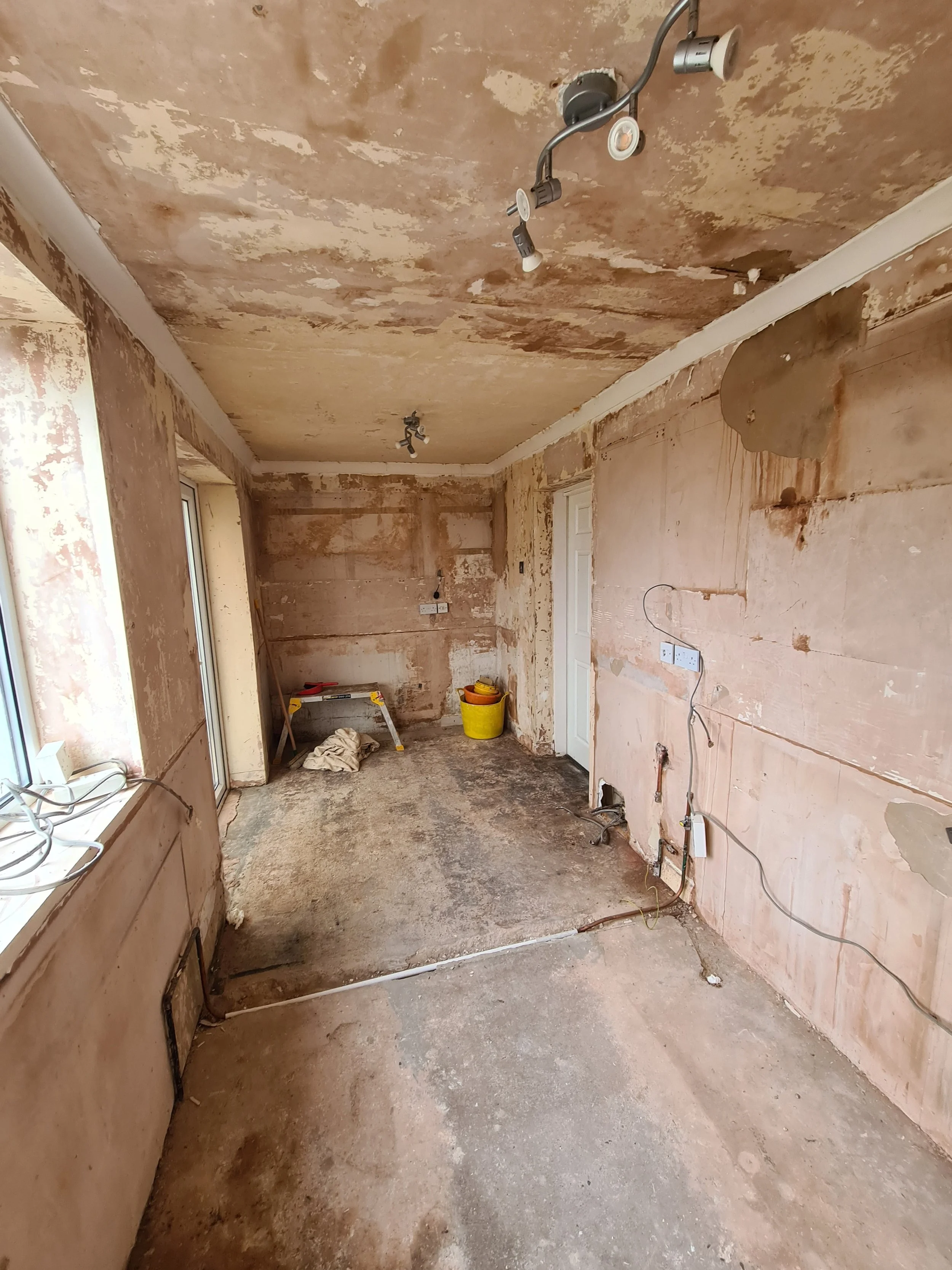 A room under renovation with stripped walls and ceiling, exposed wiring, and construction tools and materials on the floor.
