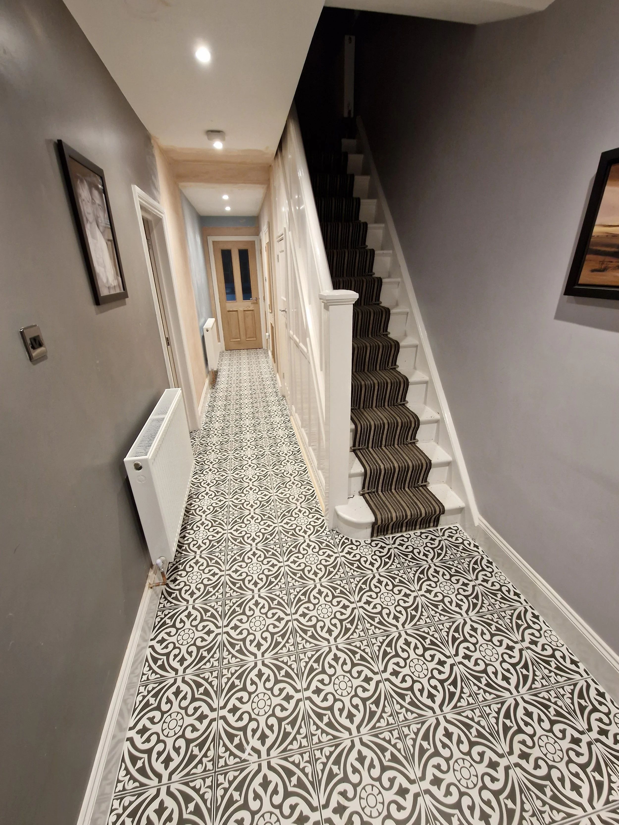 Interior hallway with patterned black, white, and gray tiled floor, gray walls, white baseboards, radiator, staircase with striped brown and black runner, and closed wooden door at the end.