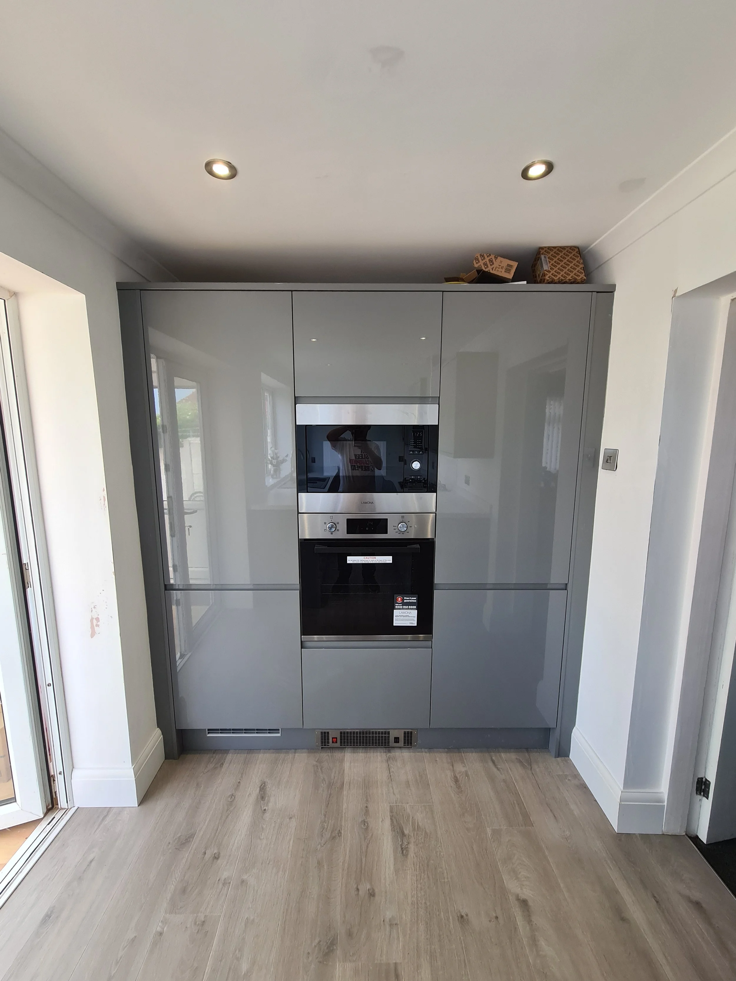 Modern kitchen with built-in oven and microwave, grey cabinetry, light wood floor, and white walls, with two small ceiling lights and a window reflection.