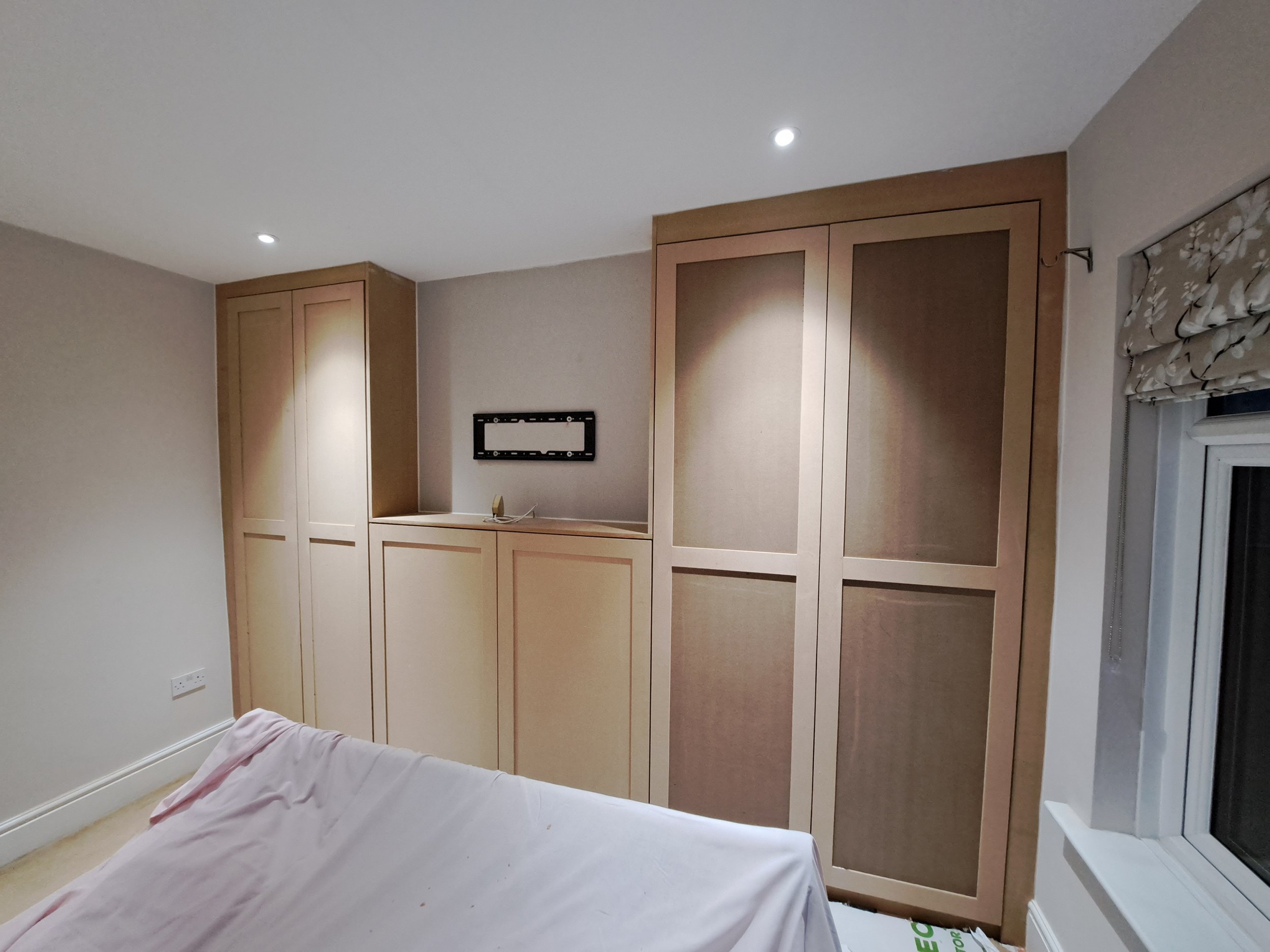 Bedroom with built-in wooden wardrobe, a window with a floral Roman blind, part of a bed with a white sheet, and a blank picture frame on the wall.