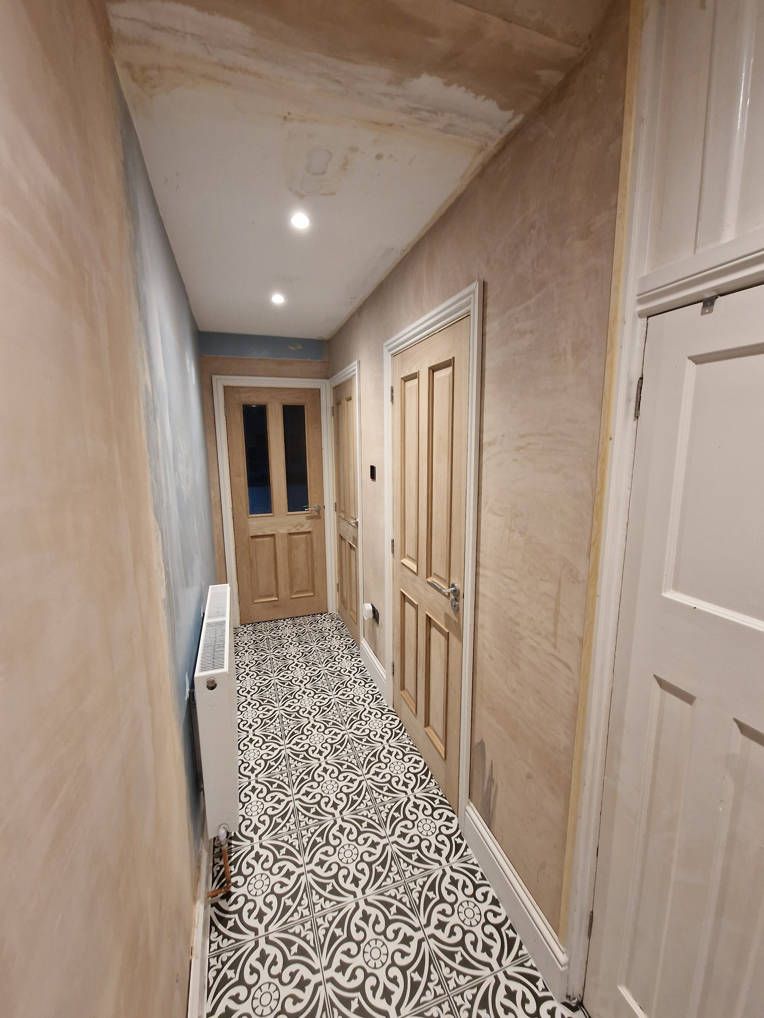 A hallway under renovation with unfinished salon walls, patterned black-and-white tile flooring, three wooden doors, and ceiling recessed lights.