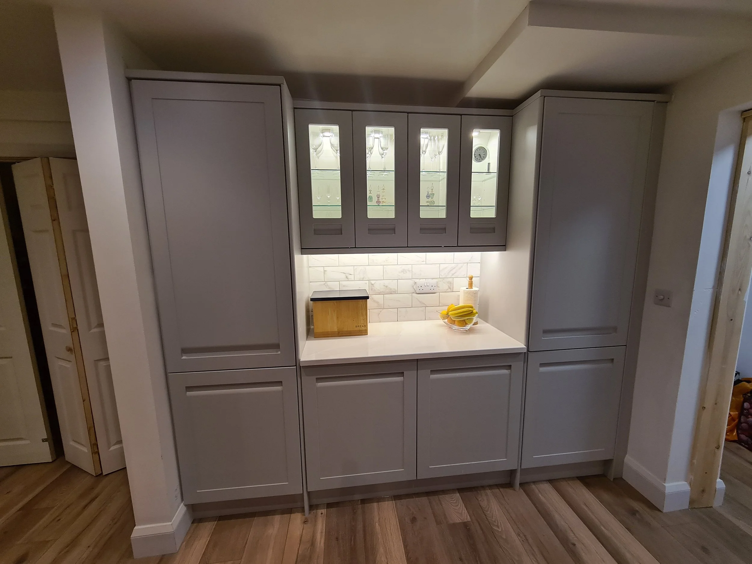 Modern kitchen cabinet with glass fronted doors displaying glassware and a small clock, with a countertop holding a bowl of bananas and a bread box.