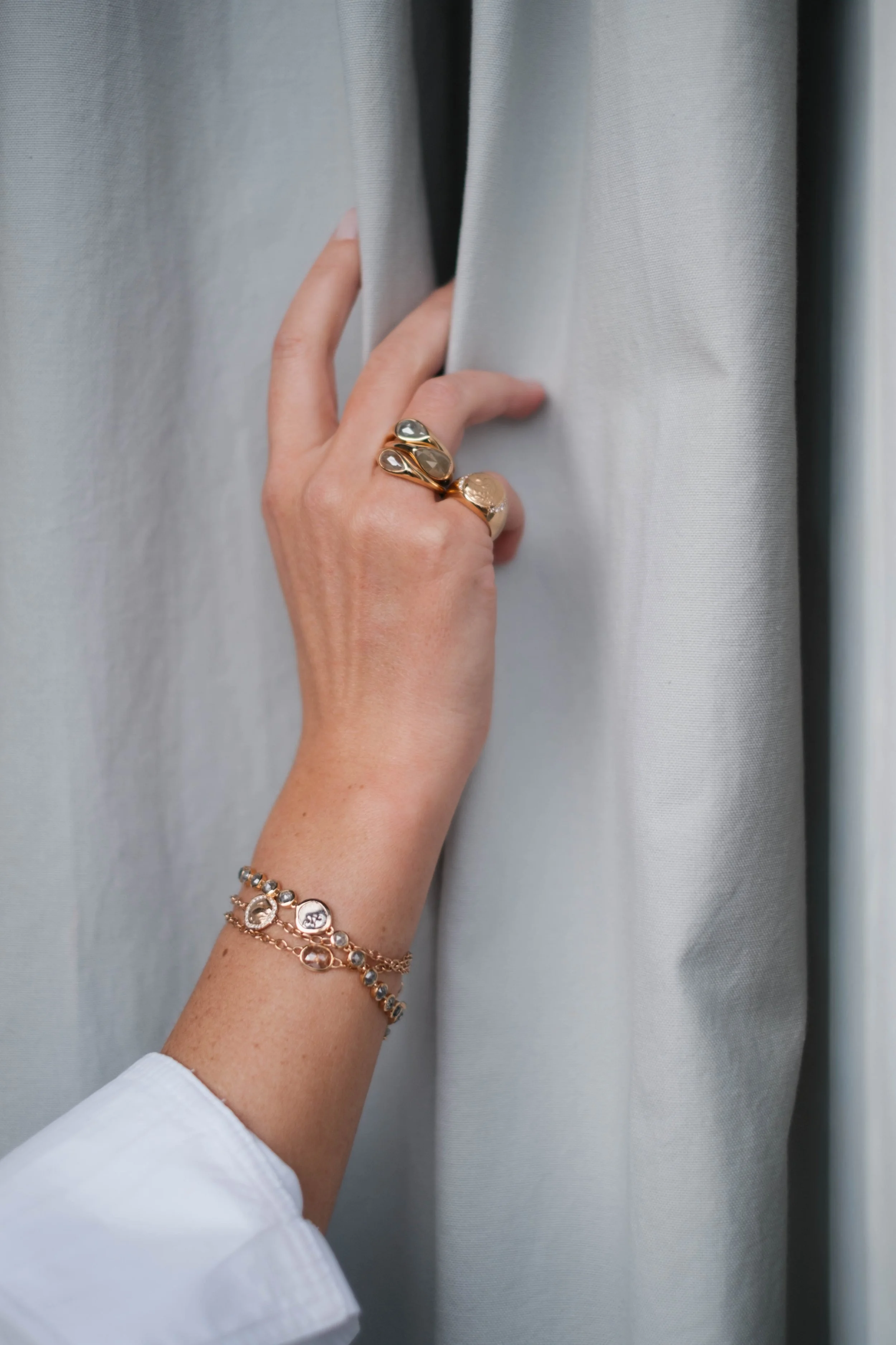 Eine Hand mit mehreren Ringen und Armbändern, die einen grauen Vorhang berührt.
