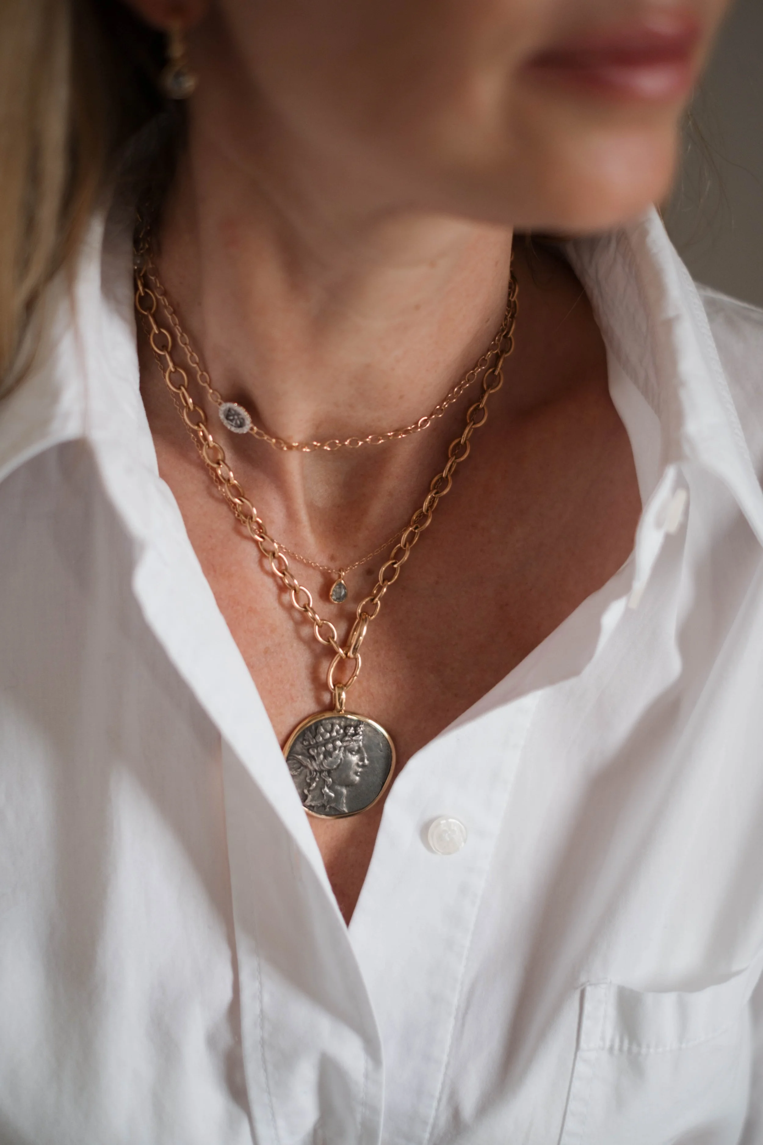 Nahaufnahme von einer gebräunten Frau, die weiße Bluse trägt, mit drei goldenen Halsketten, darunter eine große Münz-Halskette, und goldene Ohrringe.