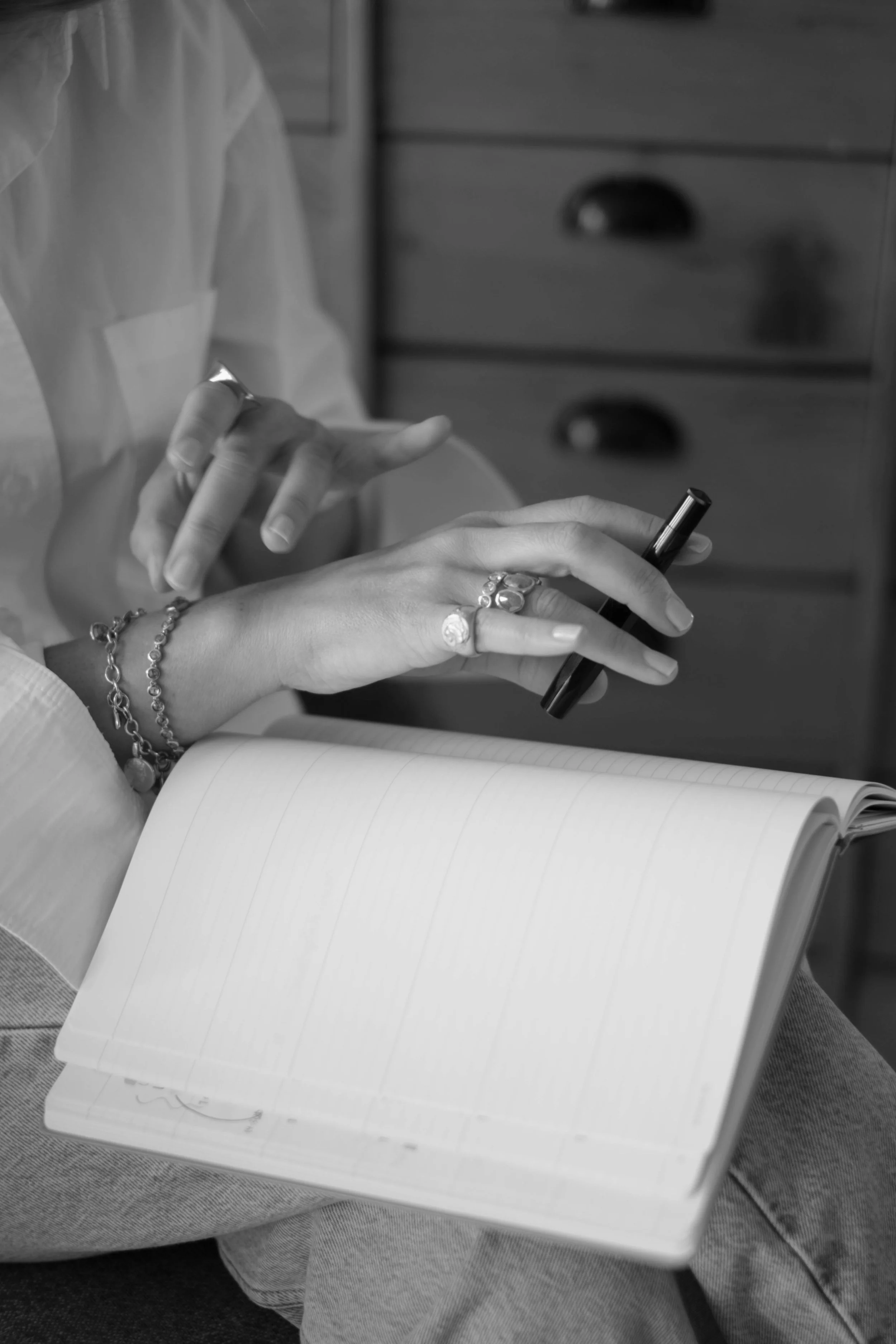 Nahaufnahme von einer Person, die in Schwarz-Weiß Notizen in ein lineares Notizbuch schreibt, während sie einen Stift in der Hand hält. Die Person trägt mehrere Ringe und Armbänder und sitzt vor einem Schrank.