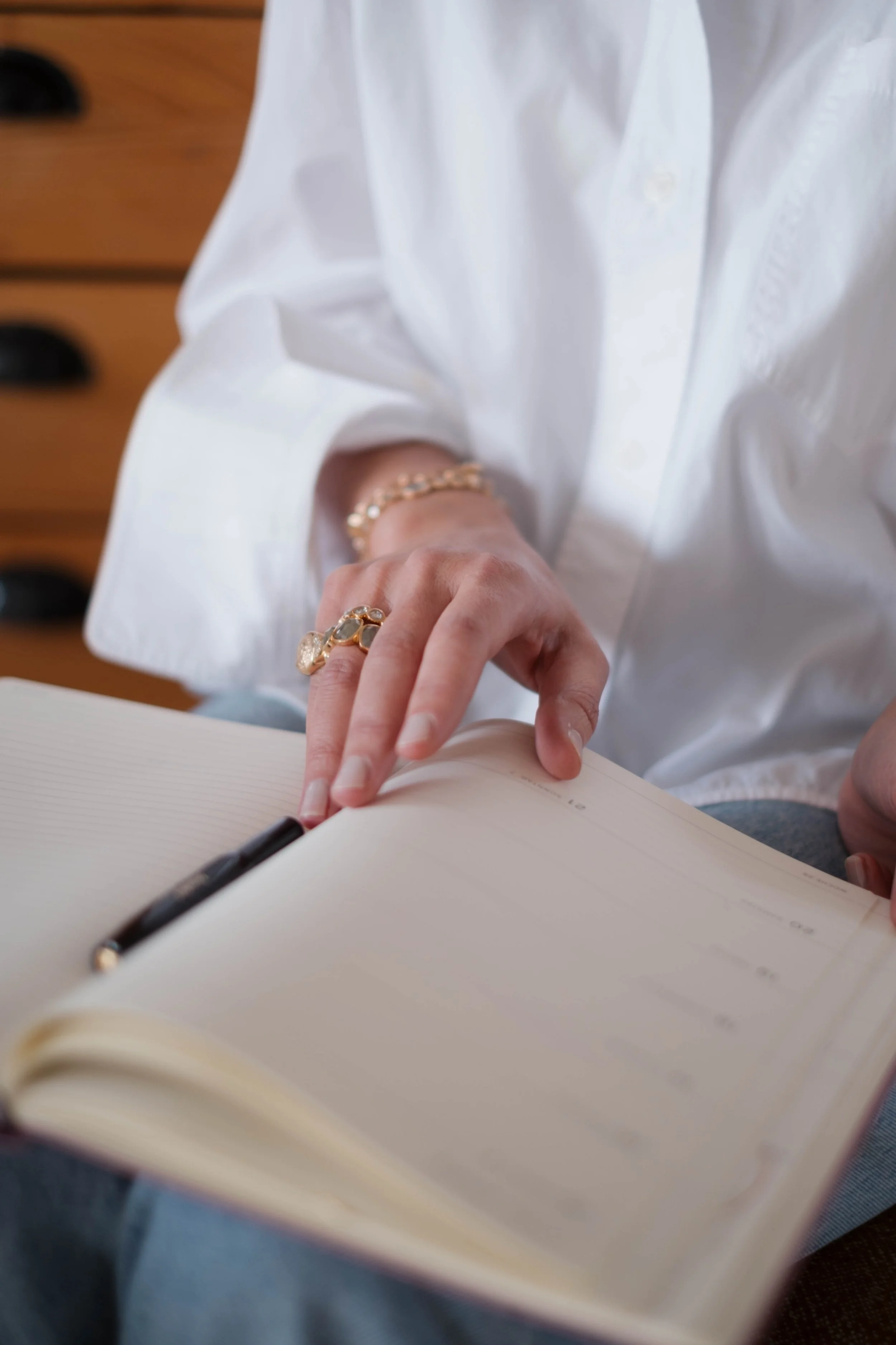 Nahaufnahme einer Person, die ein Notizbuch mit einem Kugelschreiber aufschlägt. Die Person trägt Schmuck, darunter Ringe, eine Perlenarmband und eine Kettenarmband. Die Person ist in einem weißen Hemd gekleidet, im Hintergrund ist ein Holzschrank si