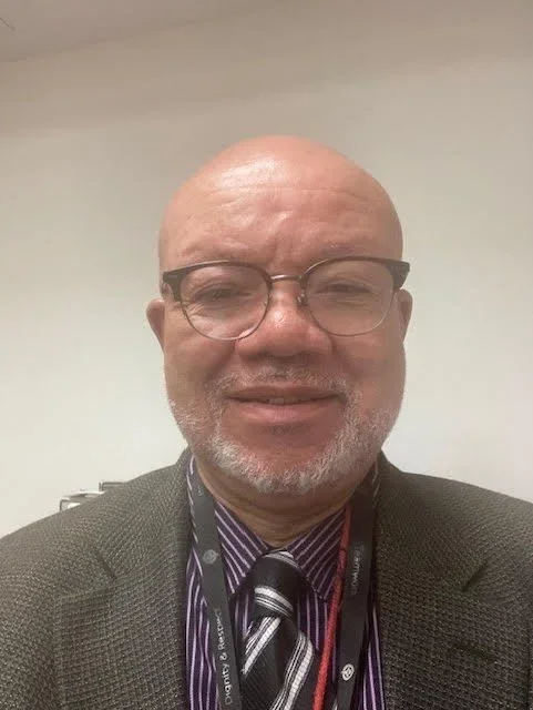 A middle-aged man with glasses and a bald head wearing a suit, tie, and lanyard, smiling in front of a plain wall.