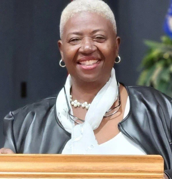 A smiling woman with short blonde hair wearing a black leather jacket, white blouse, white scarf, pearl necklace, and hoop earrings, standing behind a wooden podium.