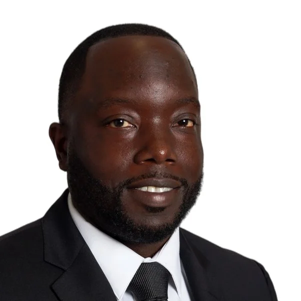 Portrait of a man with a dark complexion, wearing a black suit, white shirt, and black tie, smiling against a white background.