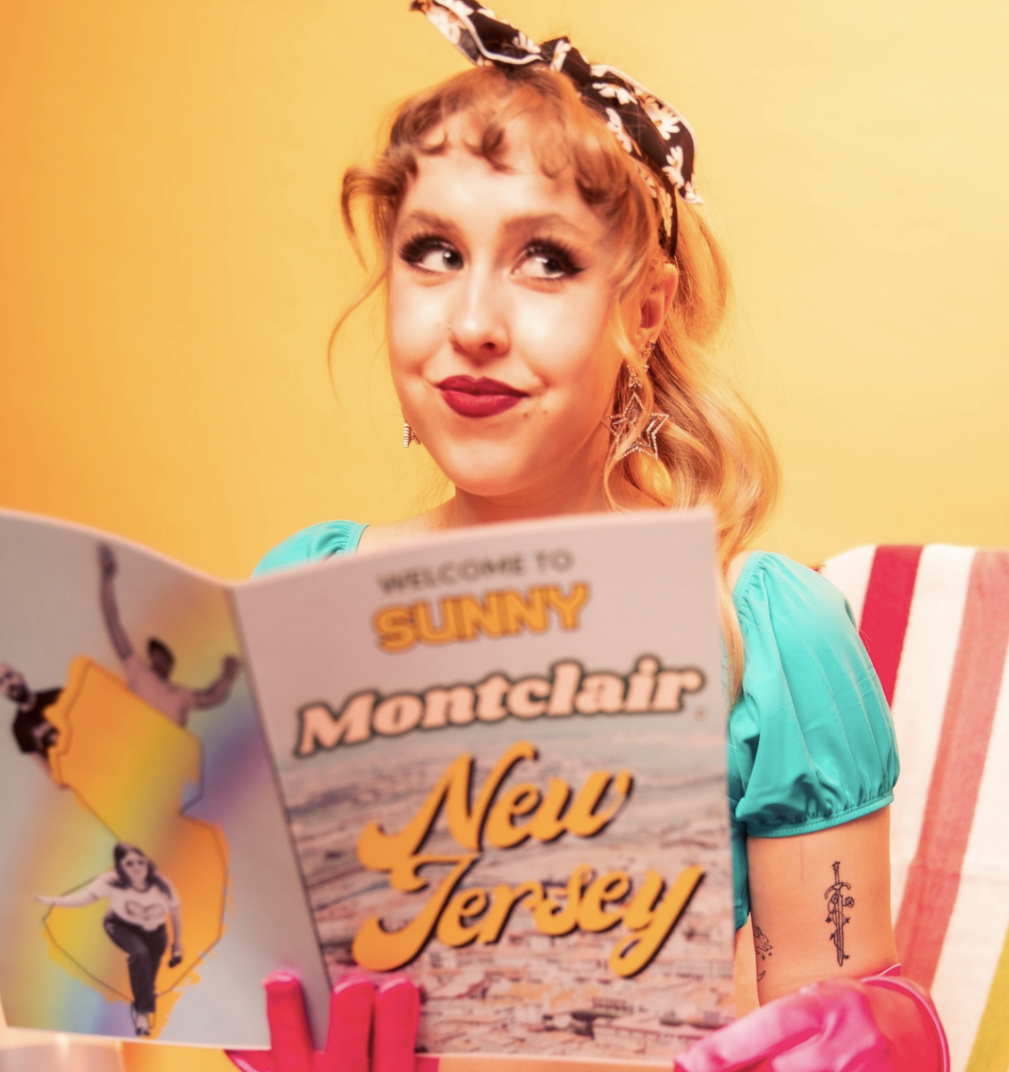 A woman with red hair and vintage fashion, wearing a blue top, holding a tourist brochure titled 'Welcome to Sunny Montclair, New Jersey.' She has a playful expression, with a slightly raised eyebrow and lips puckered, and is wearing a headscarf, sta