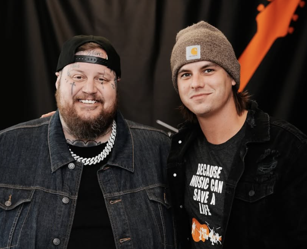 Two men smiling, one with tattoos and a black cap, wearing a denim jacket, and the other wearing a brown beanie and a T-shirt with music-themed text.