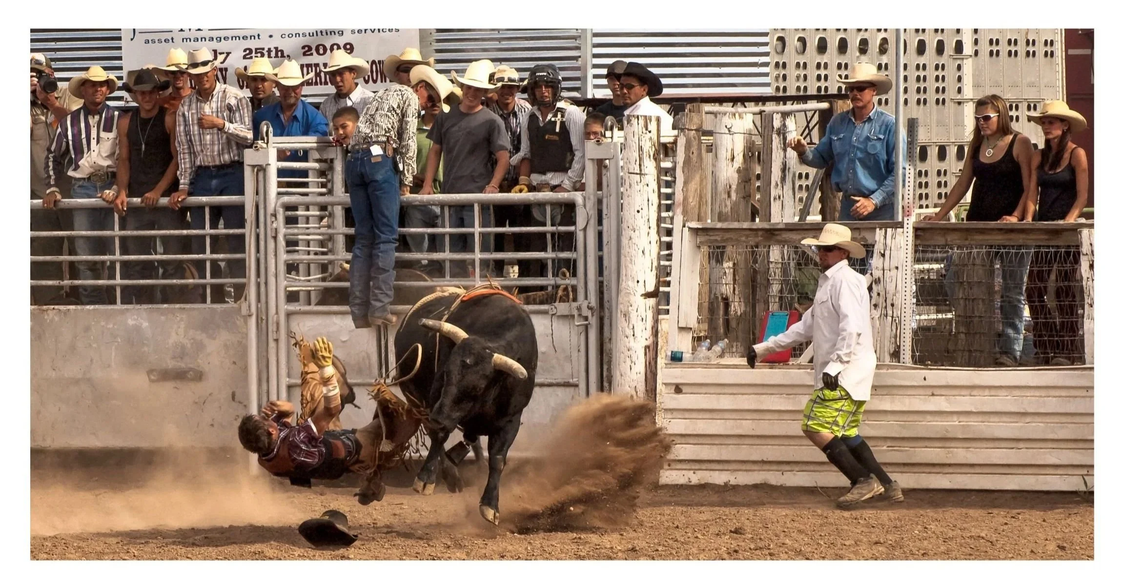 Photography of bull riding. Man falling off bull another in white coat running toward it, with people in cowboy hats watching.