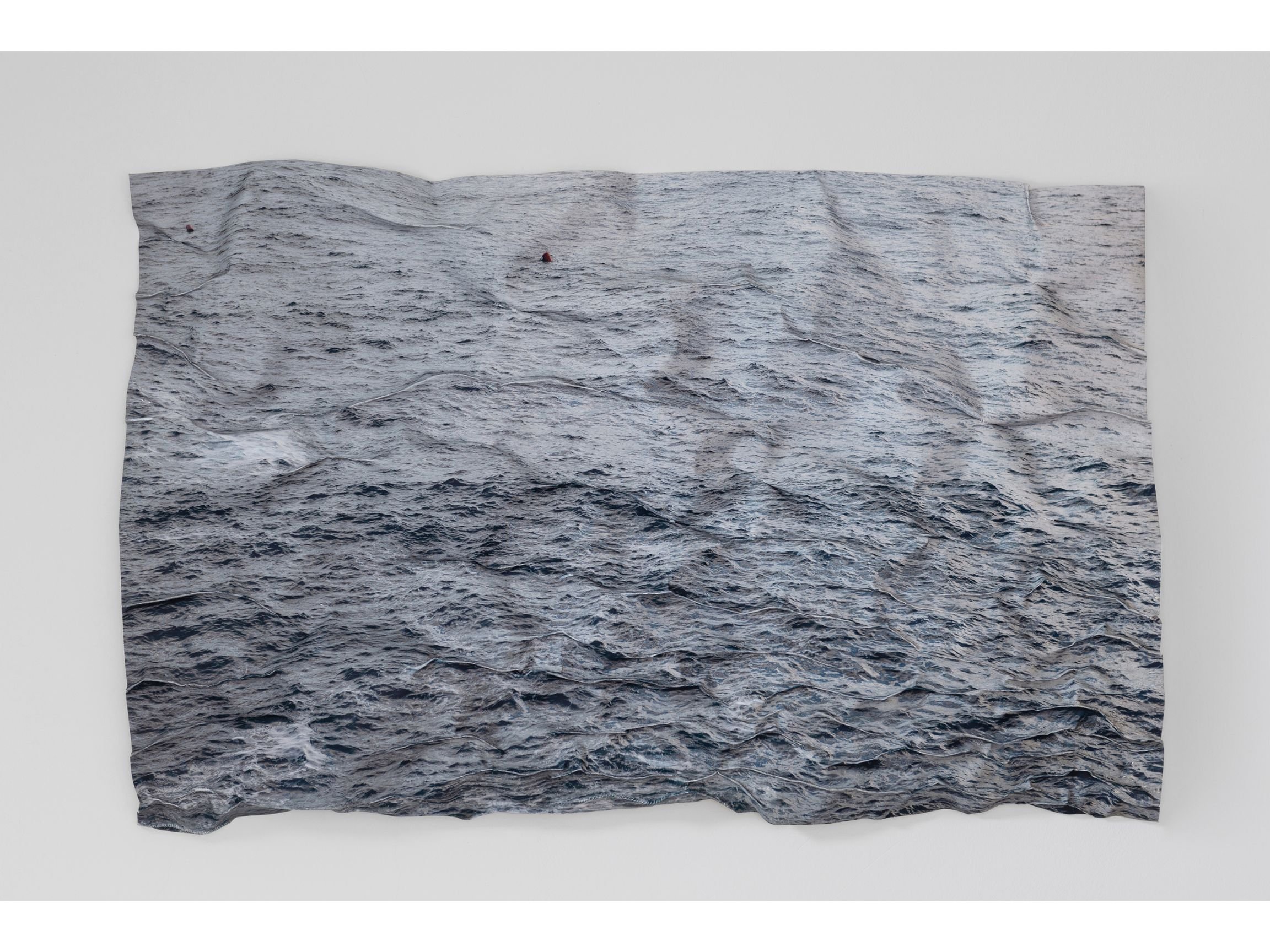Photographic artwork depicting a wide expanse of dark, choppy ocean water, printed on a textured surface with visible creases and irregular edges, mounted against a white wall.