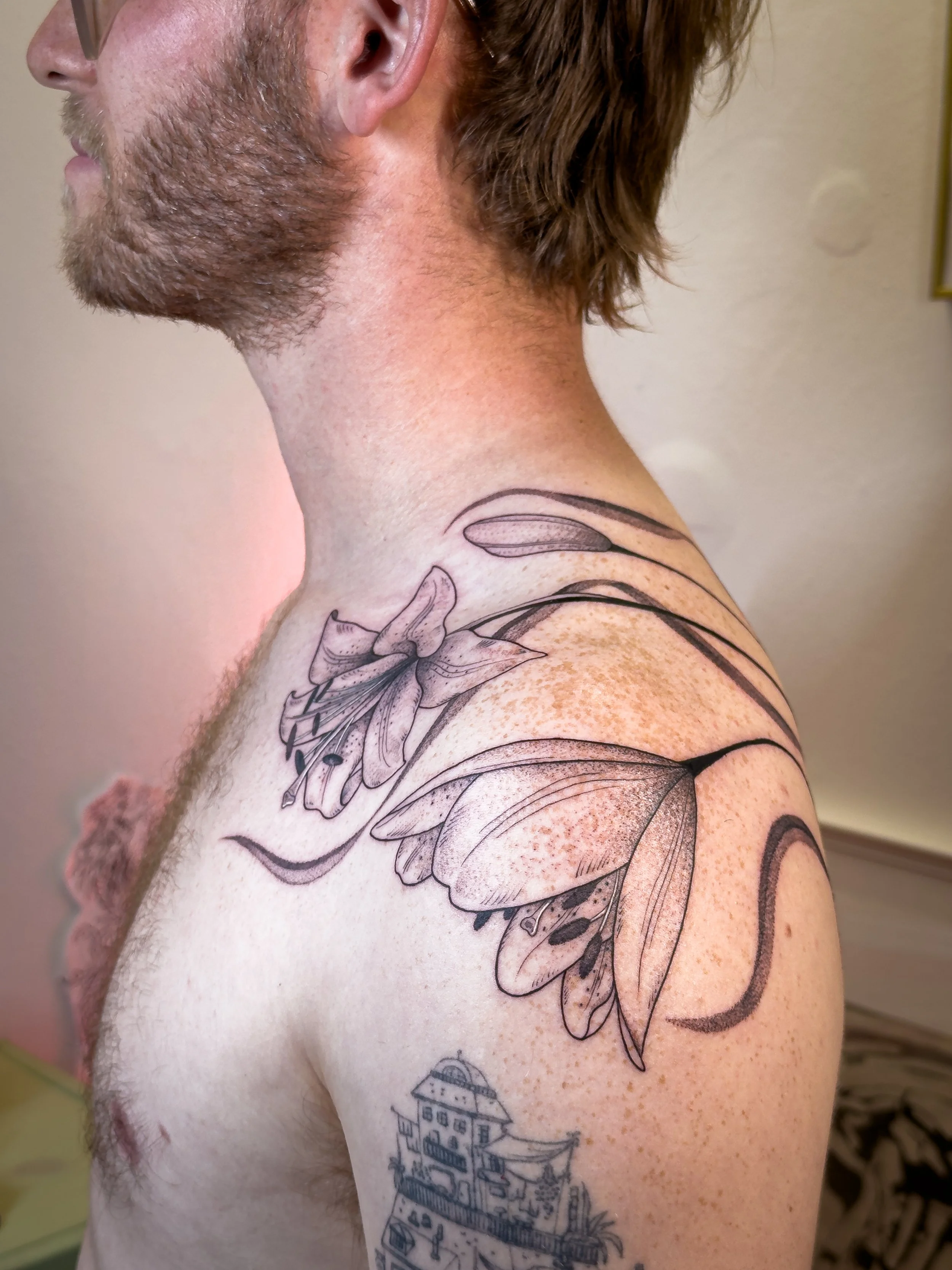 Freehand shoulder and back tattoo by Lis Roldan, featuring lily flowers, custom-designed to follow the natural movement and anatomy of the body, creating a fluid and harmonious composition that flows seamlessly with the form.