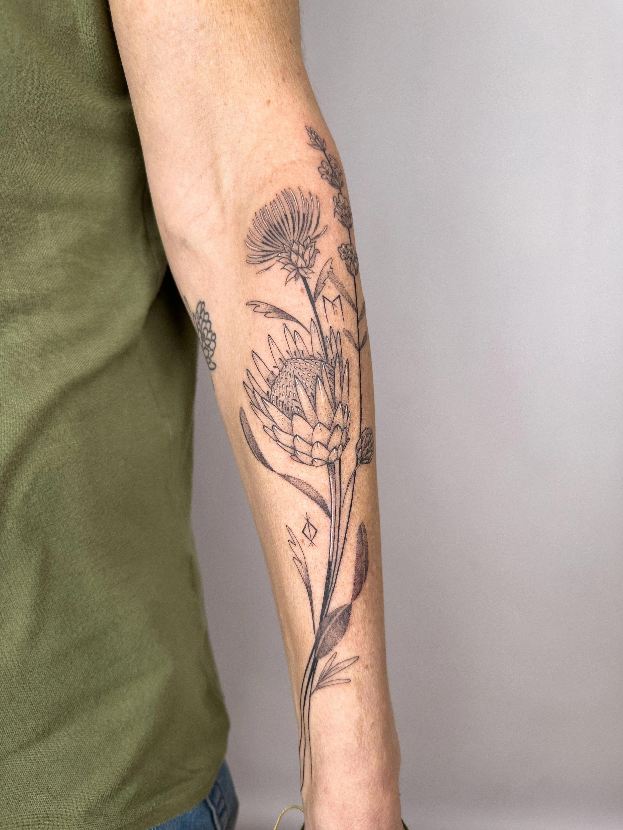 Fine line botanical tattoo featuring protea and thistle flowers, designed to flow naturally along the forearm. Soft shading and organic lines create a delicate and balanced composition.

Custom tattoo by Lis Roldan.
