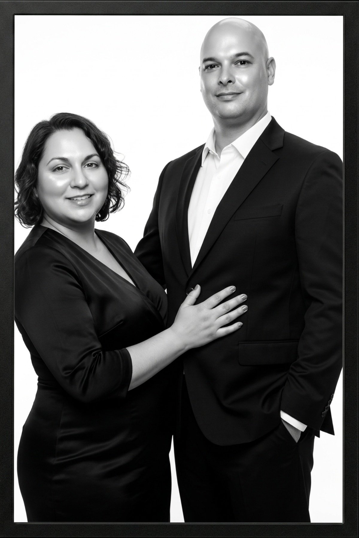 A black and white portrait of a man and woman dressed in formal attire, standing close together; she has her hand on his chest and both are smiling softly.