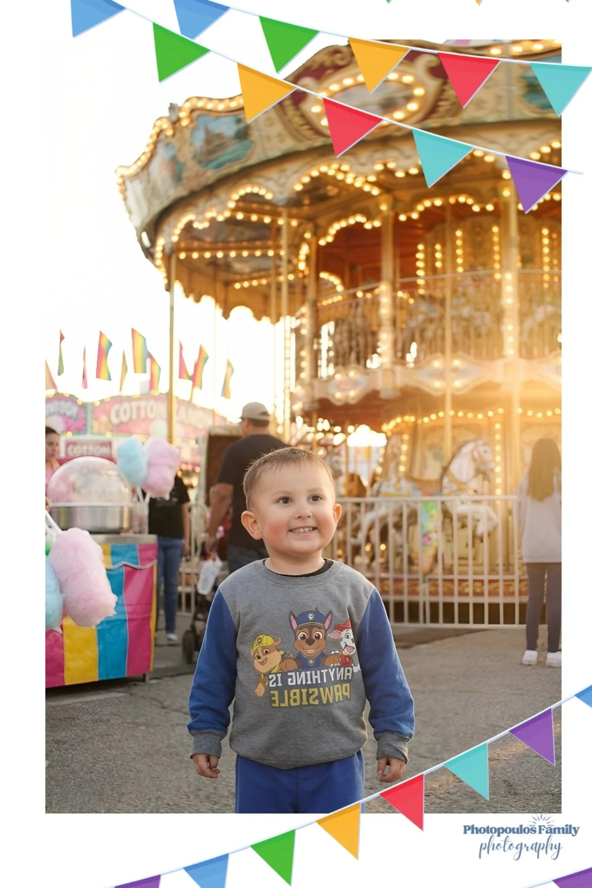 A young boy smiling at a carnival with a carousel in the background and colorful bunting overhead.