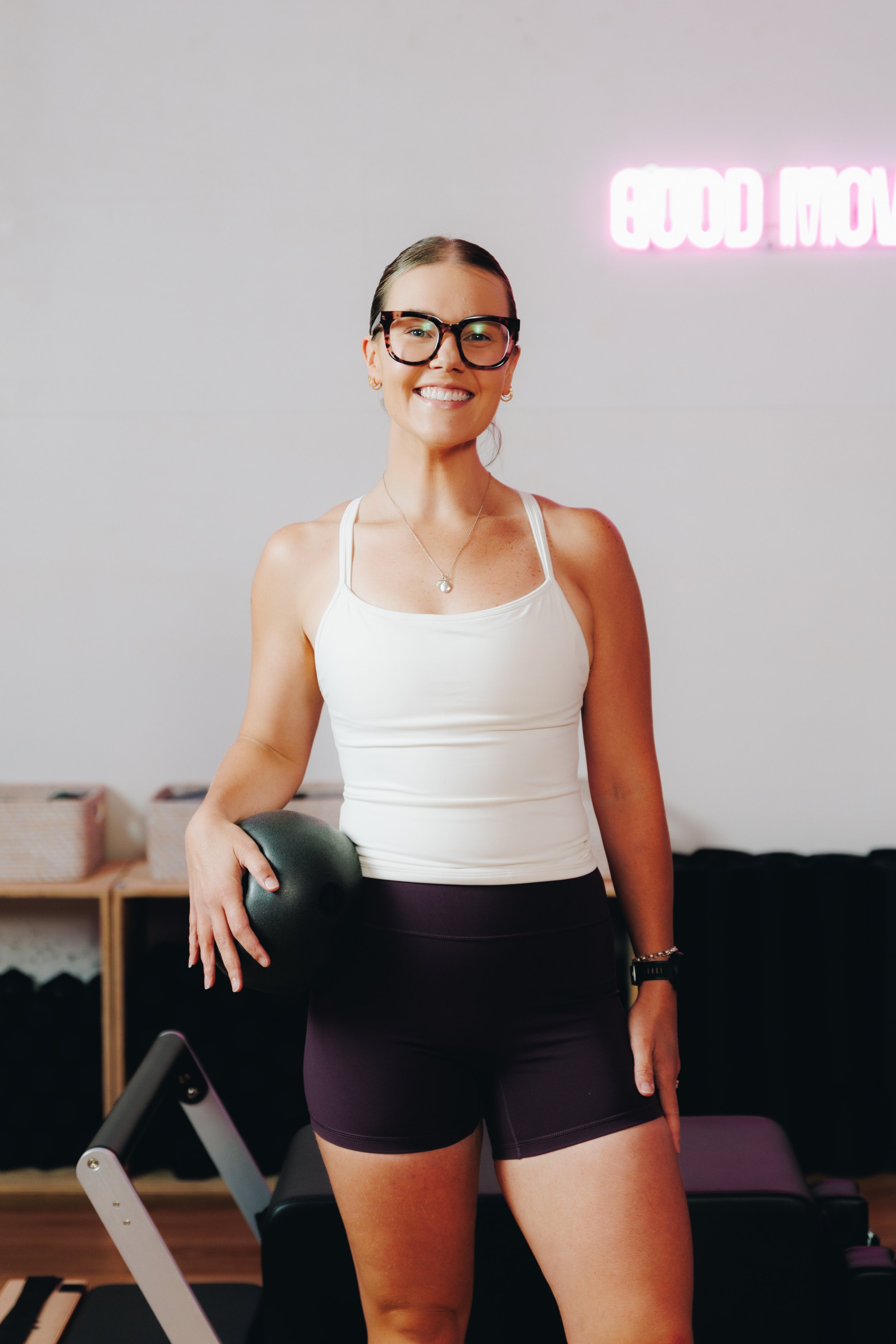 A woman with glasses smiling while holding a black kettlebell in a fitness studio with a neon sign that says 'Good Move' in the background.