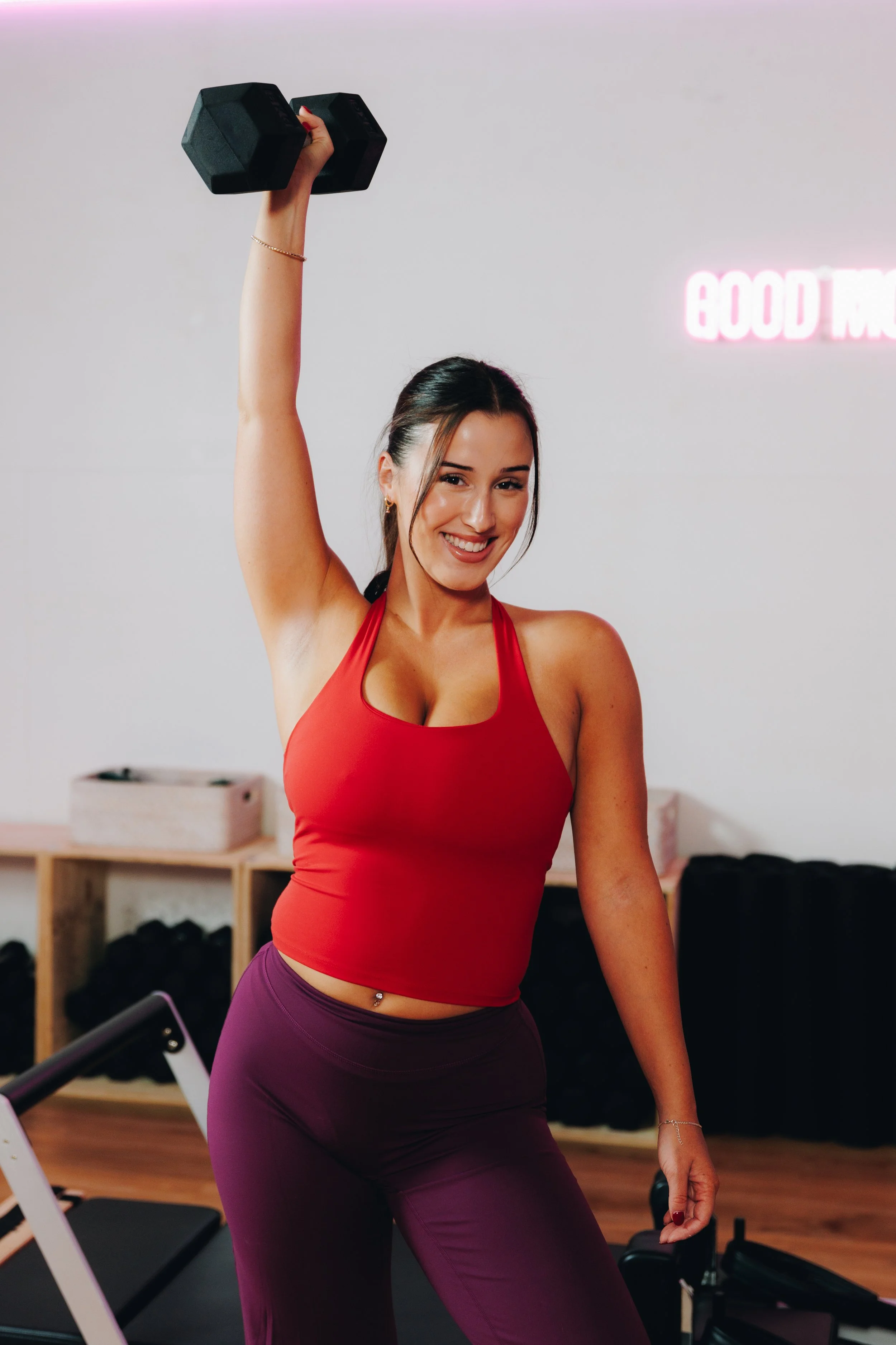 Smiling woman in red sports bra and purple leggings lifting dumbbell in gym.