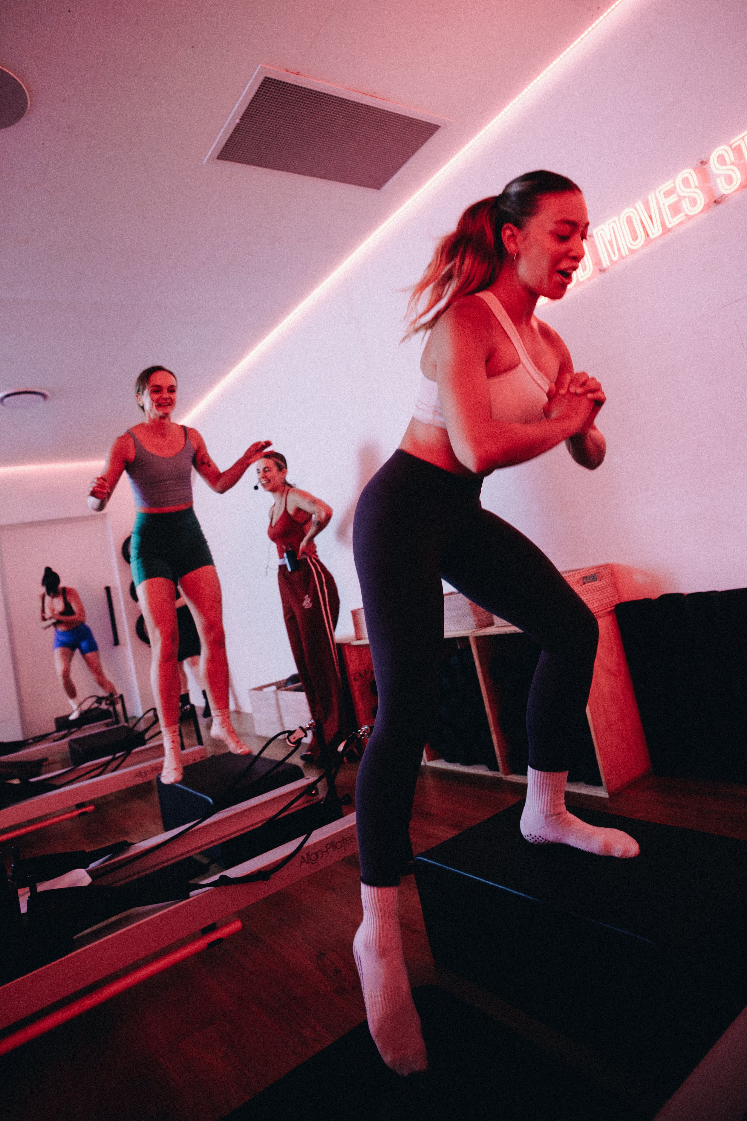 A woman exercising in a gym, leaning on a barre, smiling. There is a pink neon sign on the wall that says 'Good Moves Studio'.