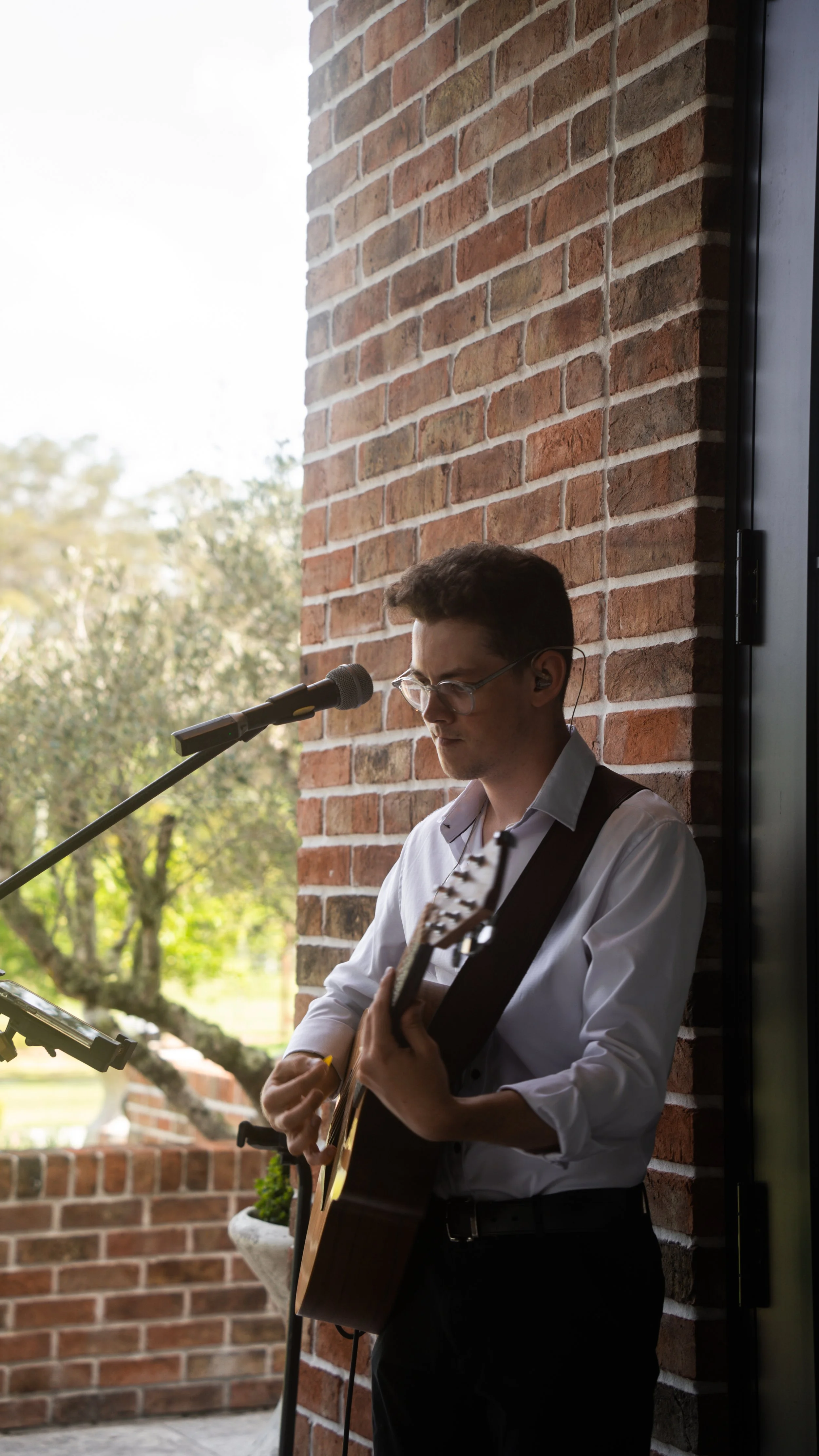 A man with glasses playing an acoustic guitar and singing into a microphone outdoors, standing next to a brick wall at a private function