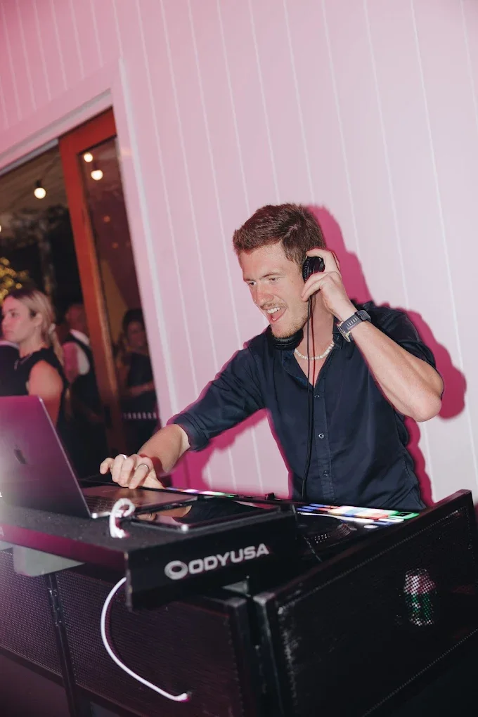 A man DJing at a wedding, wearing headphones, smiling, and using a laptop and DJ equipment.