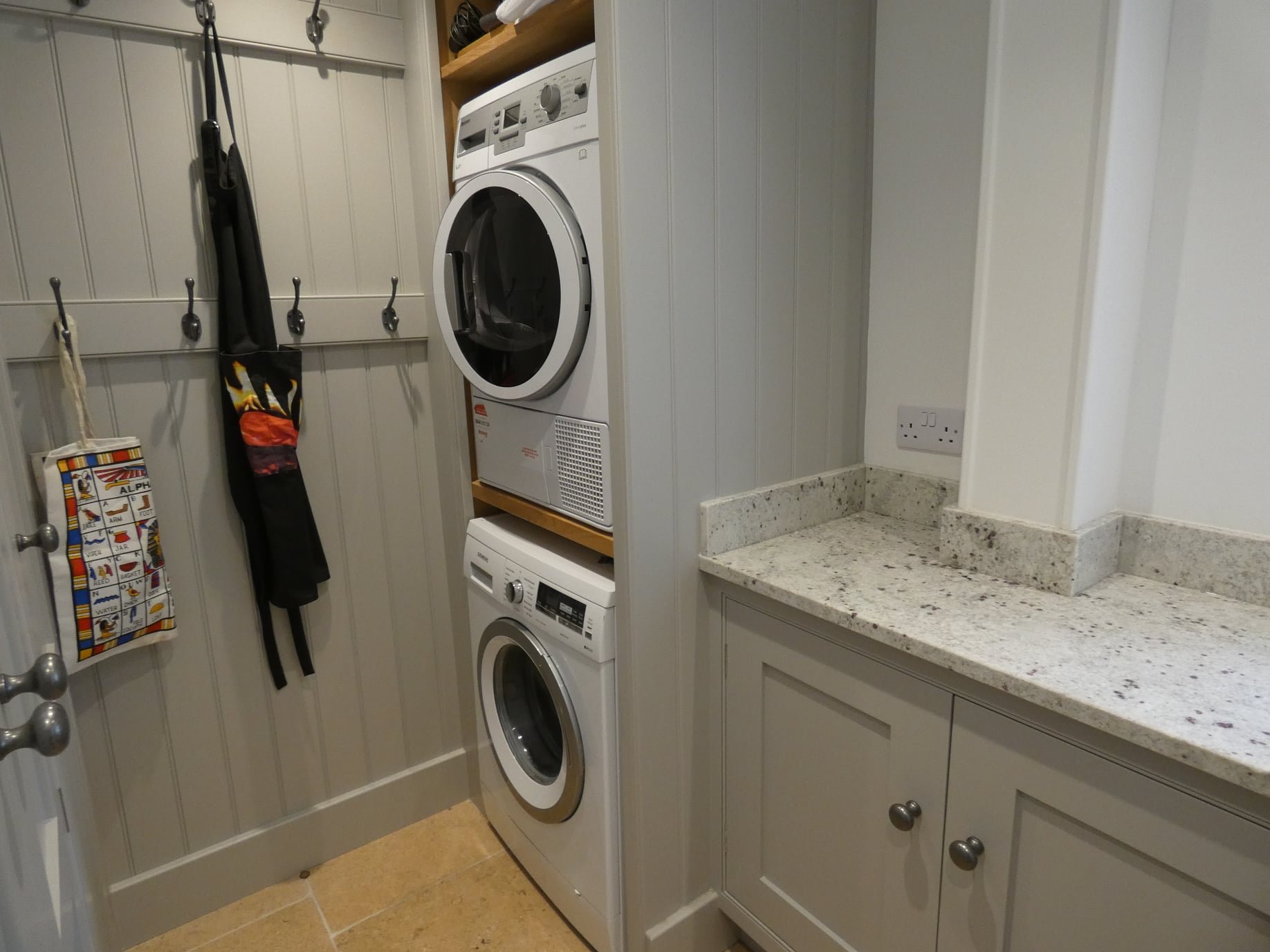 A laundry room with stacked washer and dryer units, a granite countertop, and wall hooks with accessories hanging.