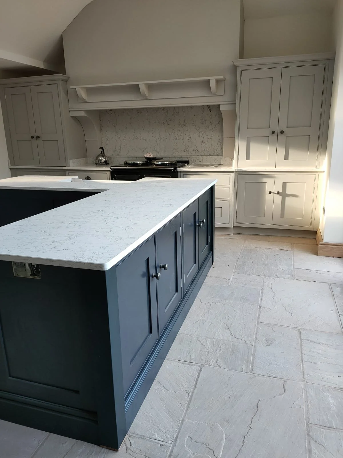 A modern kitchen with white cabinets, a white countertop, and a central island painted dark blue. There is a stove and utensils on the countertop, with beige tile flooring.