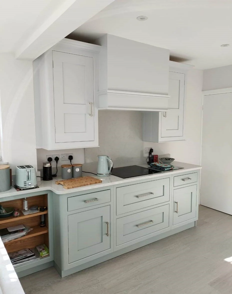 Light-colored kitchen countertop with appliances, cutting board, and wooden shelves underneath, against white cabinetry and wall, with a closed white door to the right.