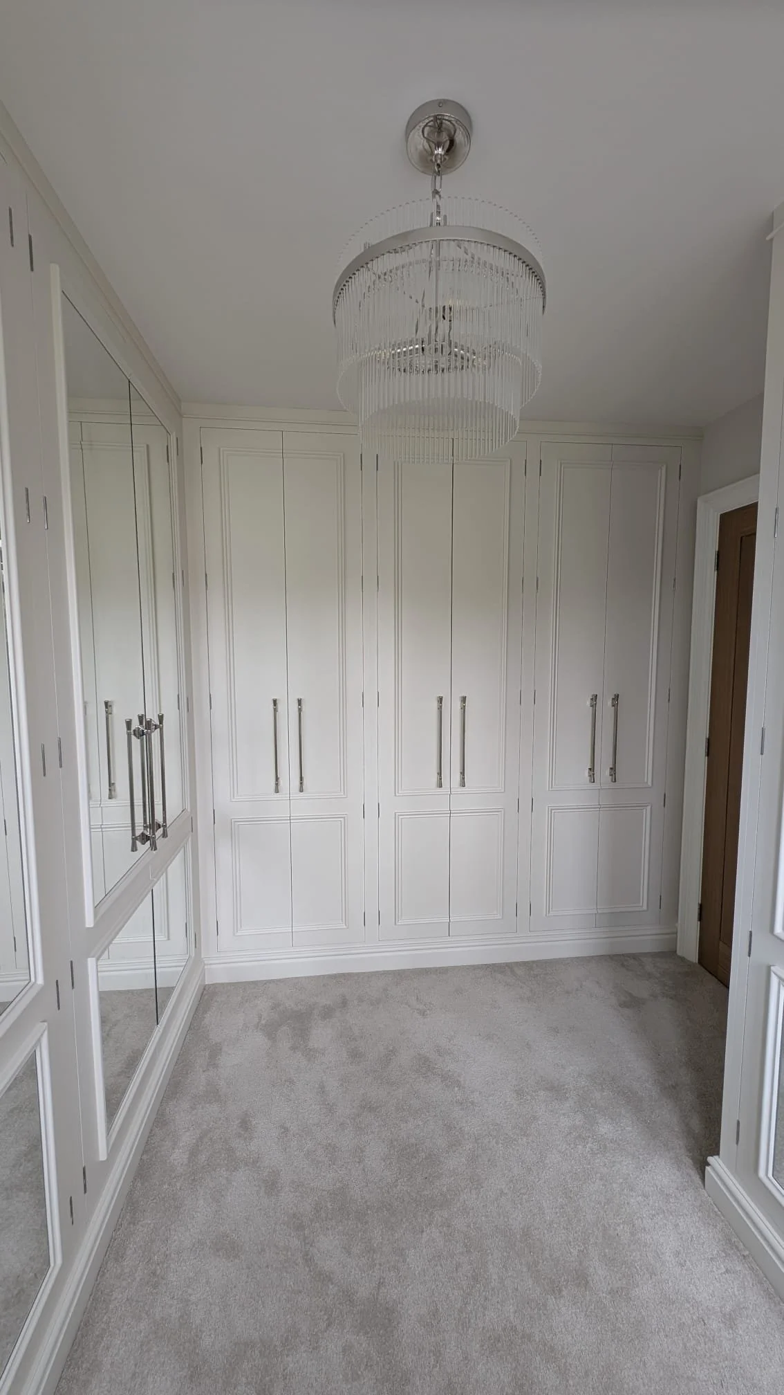 A walk-in closet with white cabinets, mirrored closet doors on the left, and a chandelier hanging from the ceiling.