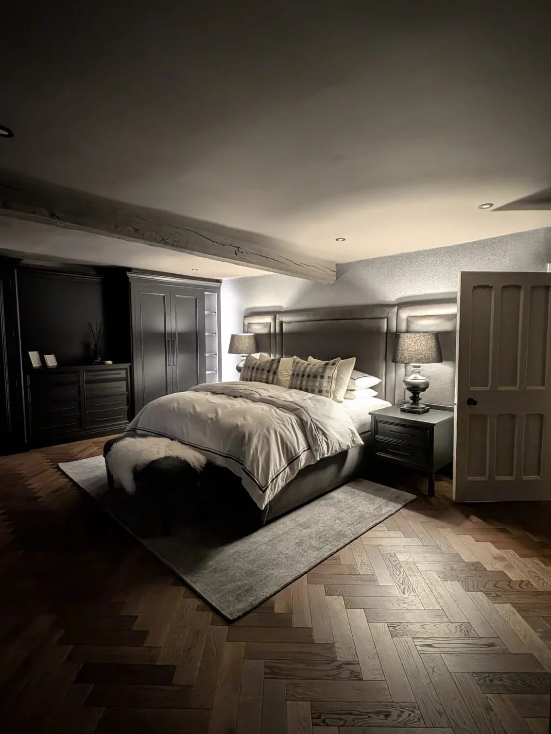 A cozy bedroom with a large bed, two nightstands with lamps, and a dark wardrobe, on a hardwood floor with a small rug.